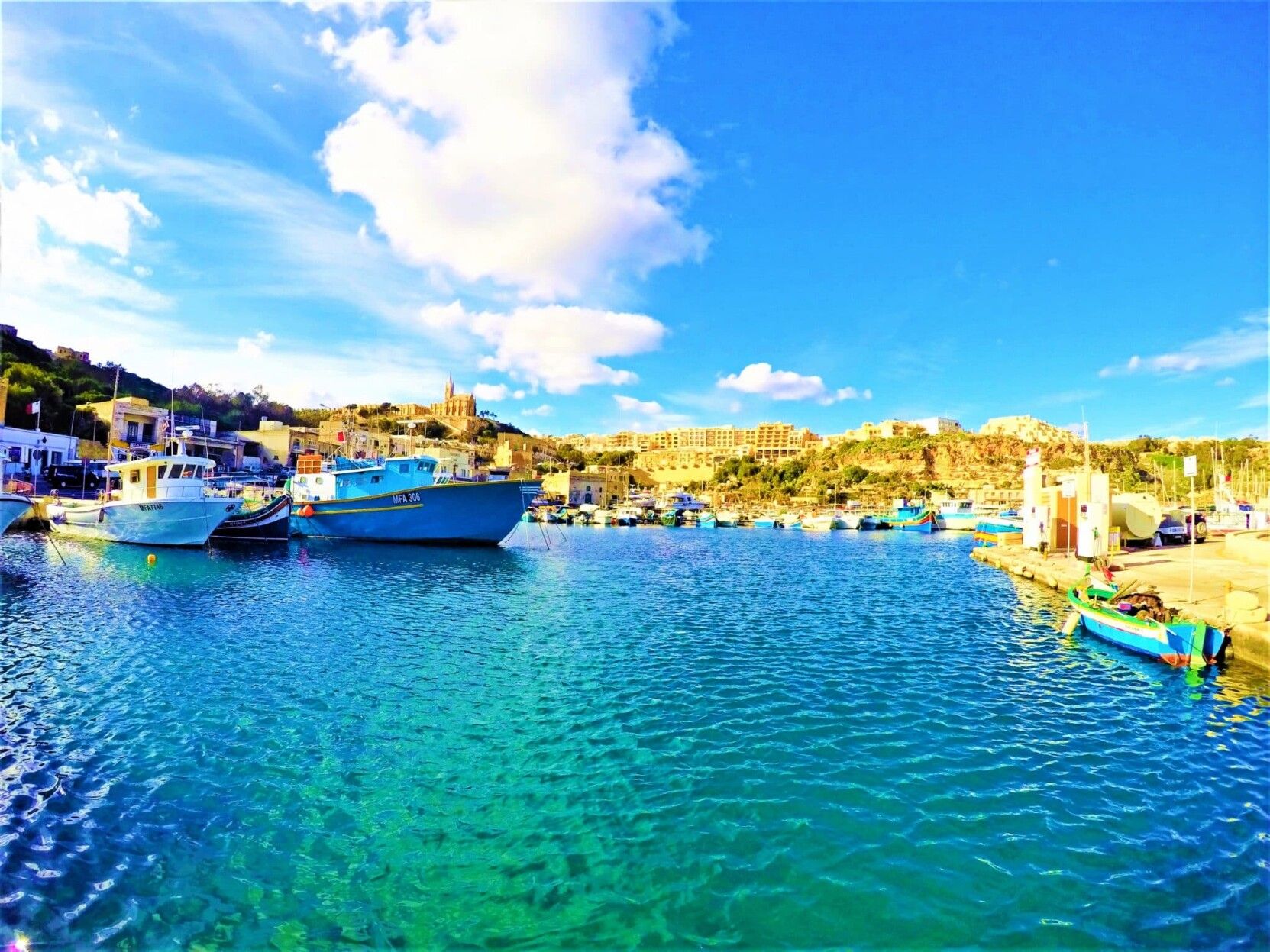 The image shows a bay in the south of Malta. The Mediterranean sea a crystal blue, clear and clean, still, with the Luzzu and the Dgħajsa (The dgħajsa and luzzu are typical Maltese boats with unique features, used mostly by Maltese fishermen and frequently found in smaller harbours ) under a crisp blue sky with wisps of cloud. The sun soaks everything in a comfortable warmth.