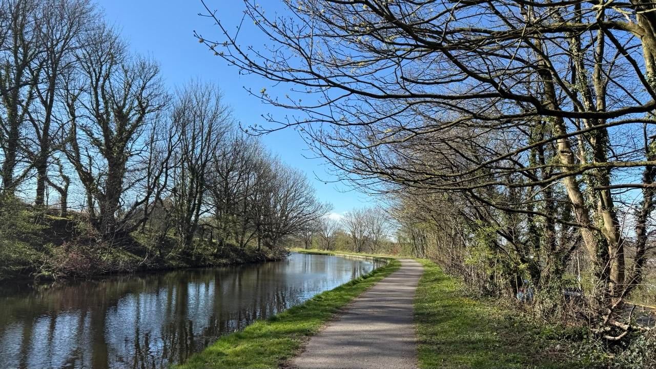 A walkway leads alongside an aqueduct. The sky is blue and the trees branches are still bare in this scene from early spring 