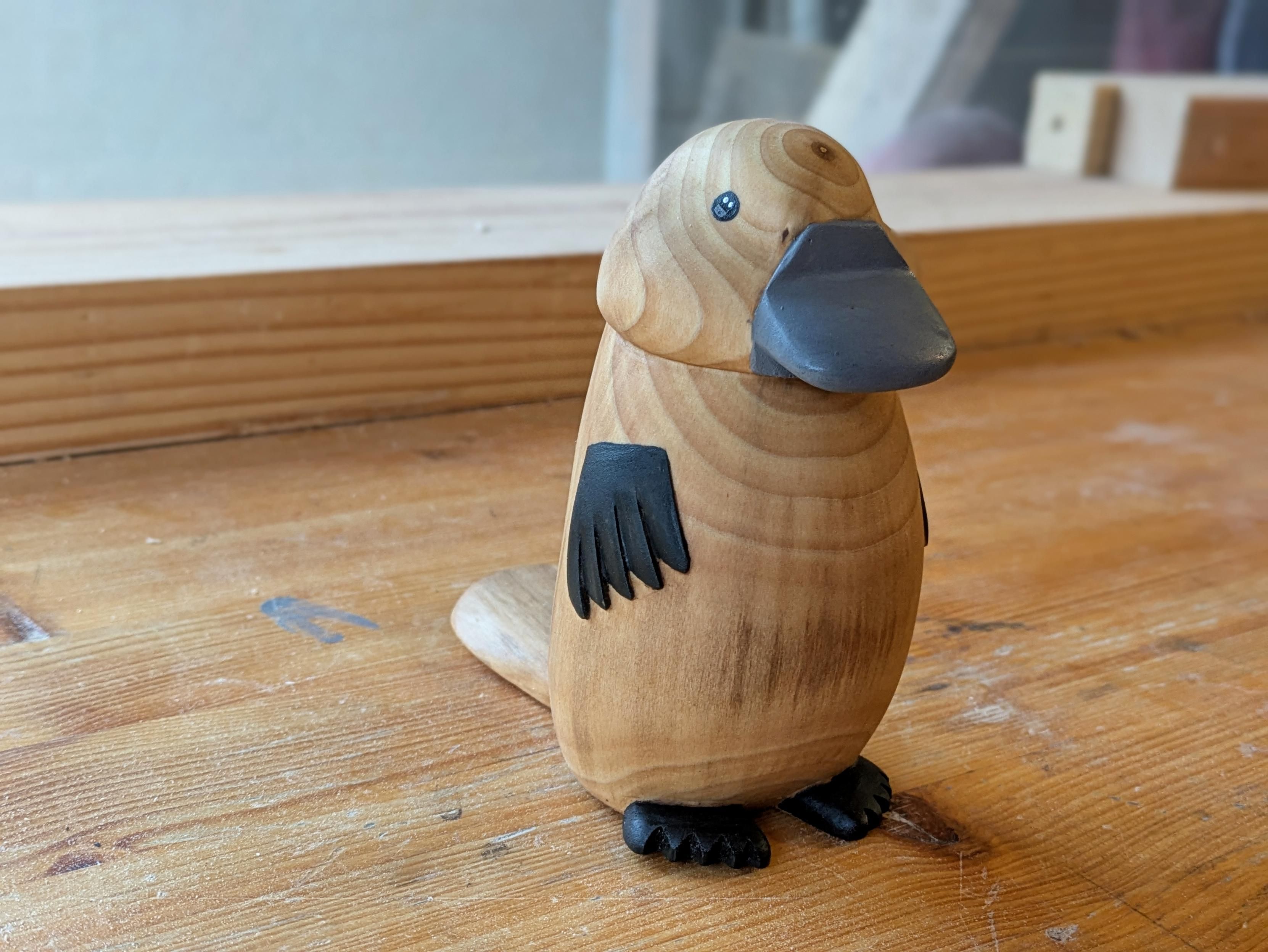 Woodcarving, sculpture of a standing up platypus. Its paws are painted in black, and its beak ing grey. It has black eyes with two white dots and a grey one.