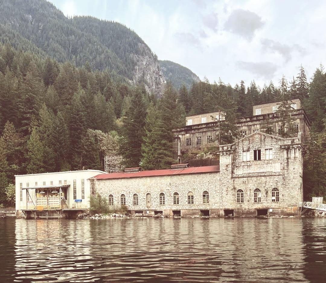 Old stone industrial building complex with arched windows, red roofs, and surrounding forest, reflected in calm water under a cloudy sky.