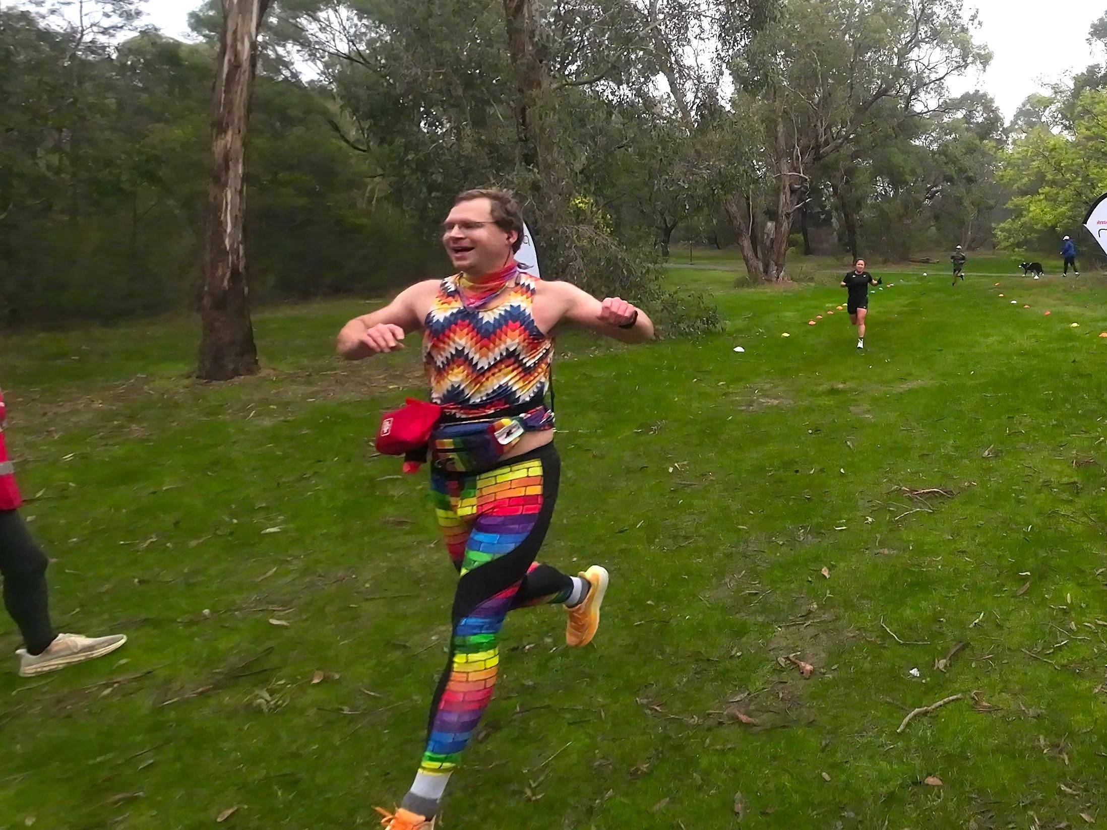 Picture of Luke just after they cross the finish line at Parkrun. They are wearing their usual Parkrun getup - pride pattern leggings, colorful top, pride colored bandanna around their neck, pride patterned hip bag plus first aid kits around their waist.

The picture was snapped mid stride, with their front foot having just landed as the back leg is bent almost at 90 degrees.

Their arms are up at shoulder height and they are smiling widely.