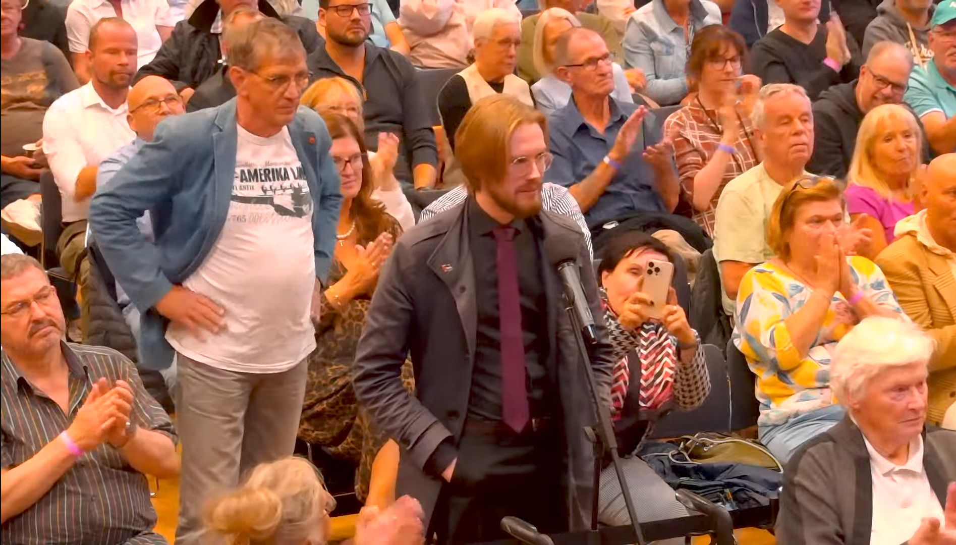 Ein Bild von Erik Uden mit Anzug, Krawatte und Palästina Pin auf einer AfD Veranstaltung. Eine Frau filmt ihn, der ganze Saal