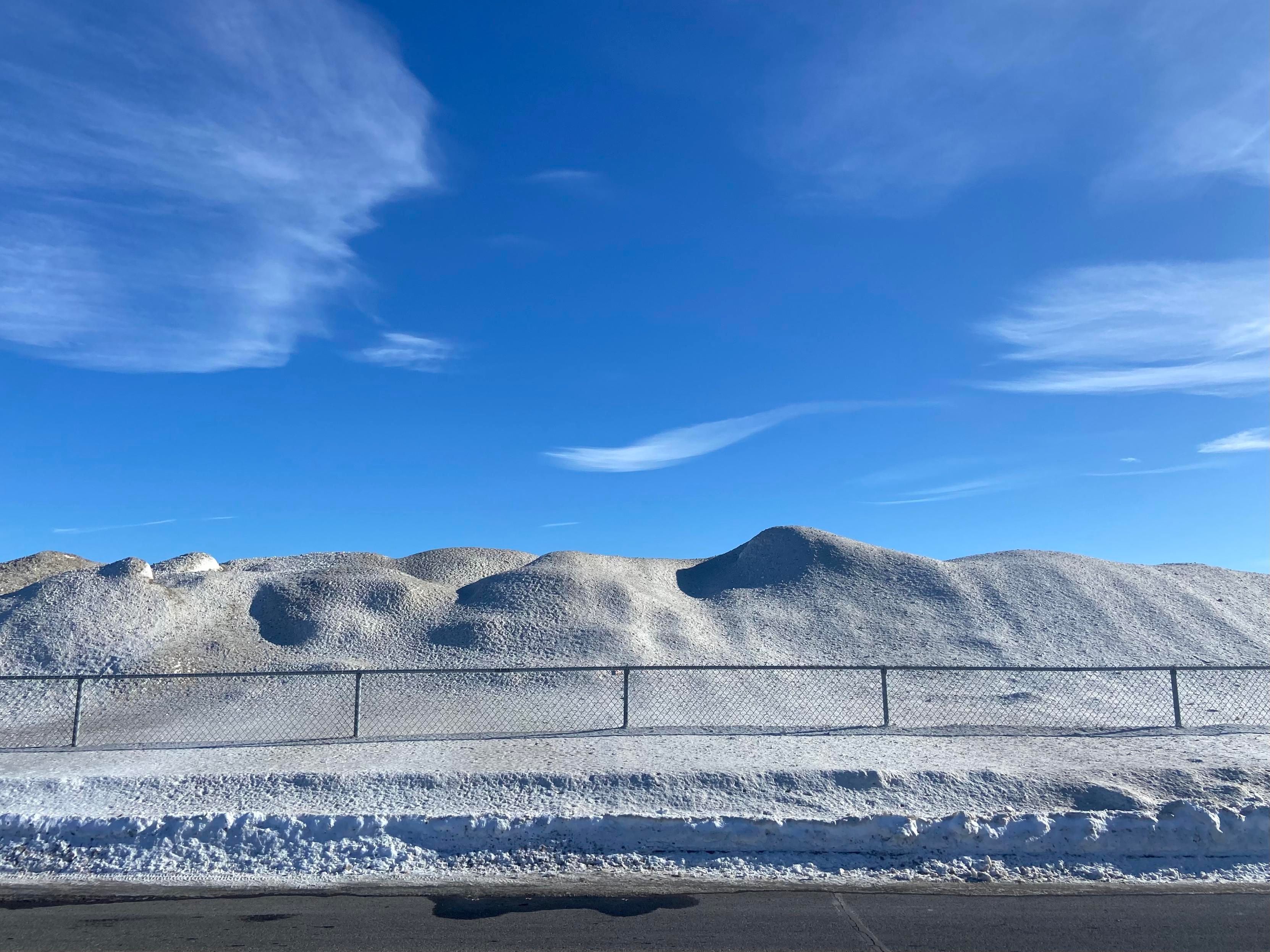 A big mountain ridge of dirty snow behind a fence. It has been removed from a parking lot. The sky is mostly blue with some wispy clouds. 
