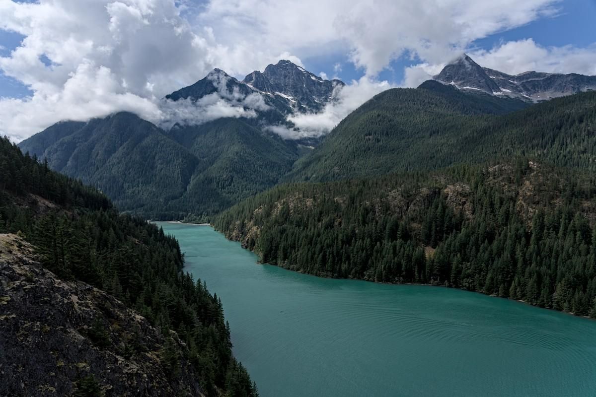 A color landscape photo of a narrow turquoise colored lake extending from the lower right to center-left. The lake is confined by steep conifer tree covered mountains. In the background is a jagged mountain range with a lot of clouds breaking up on it due to a clearing storm. There is a little bit of blue sky also visible.