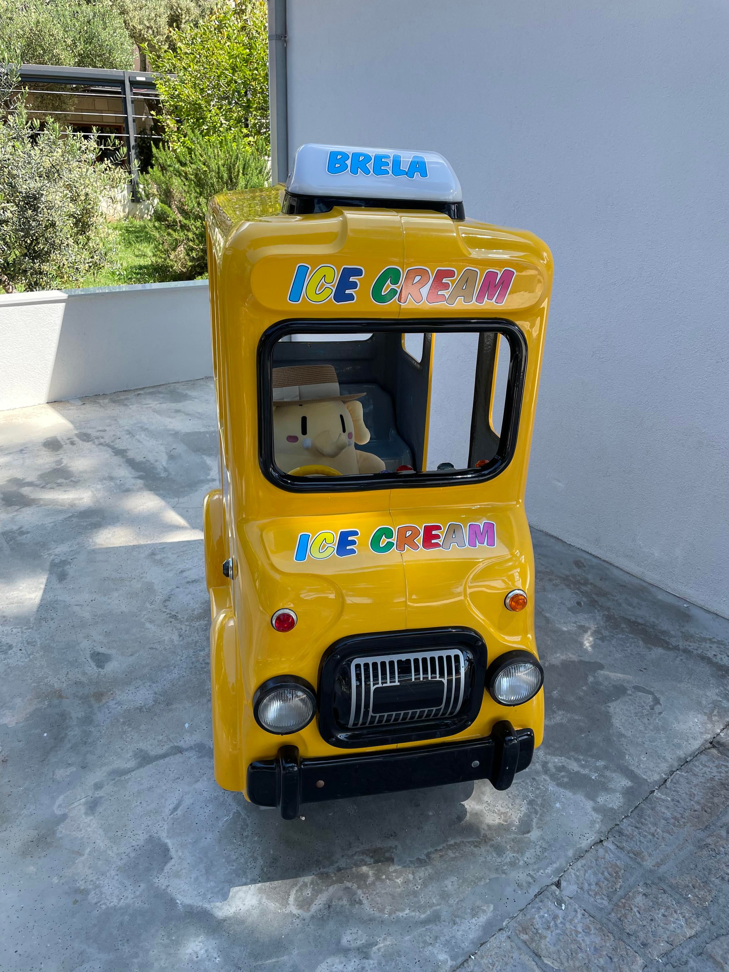 A beige stuffed Mastodon toy wearing a straw hat (not included), sitting behind the wheel of a toy ice cream truck for children (not included).