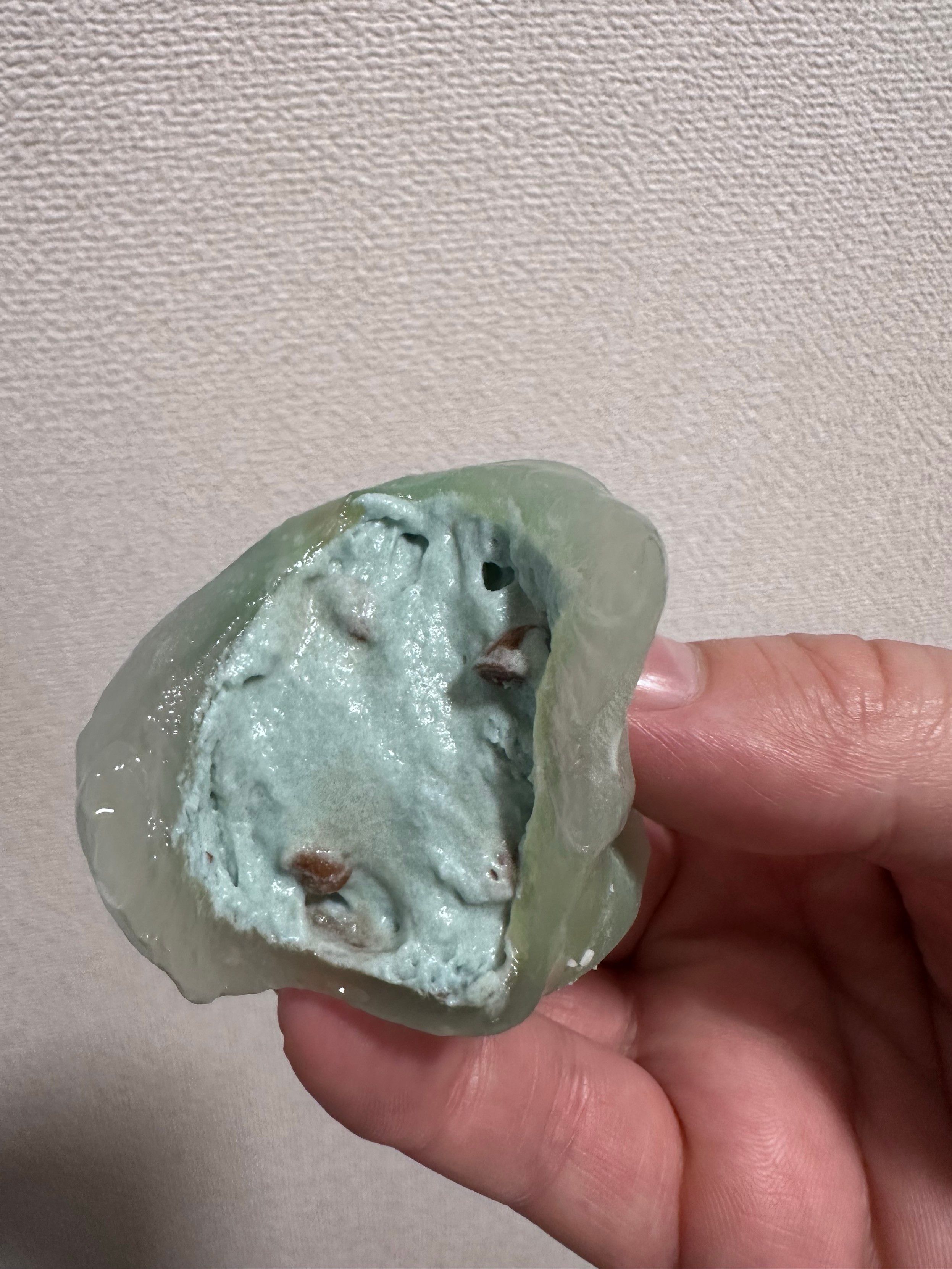 A hand is holding a piece of green jelly-like substance, revealing a pale blue interior dotted with small brown bits. The background features a light textured surface.