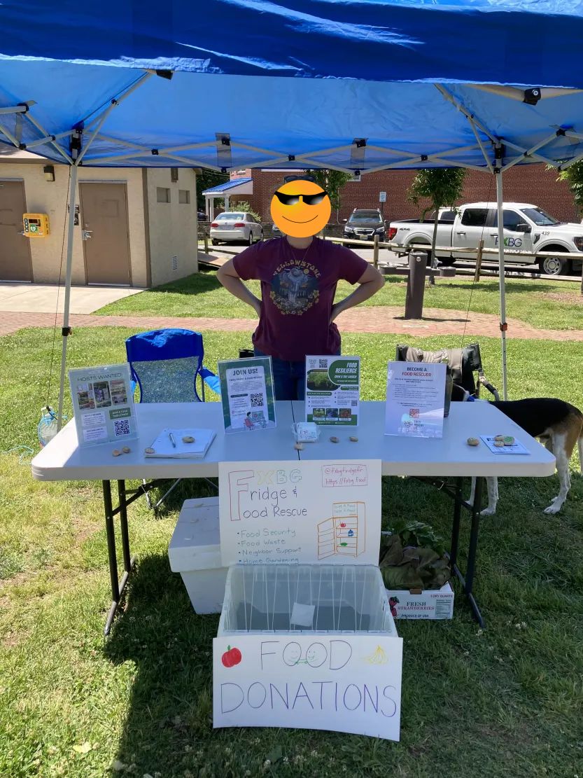 A community member mans a Fridge and Food Rescue booth at the local farmer's market. On the booth are several information placards giving info on various food initiatives. Below the booth is a bin for food donations. Standing under the shade is by the side is a friendly dog.
