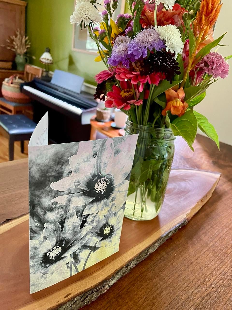 A greeting card with three blooms on the front of it, standing on a rough-hewn walnut plank. Next to the card is a clear vase filled with wildflowers. In the next room, beyond these is an upright piano.