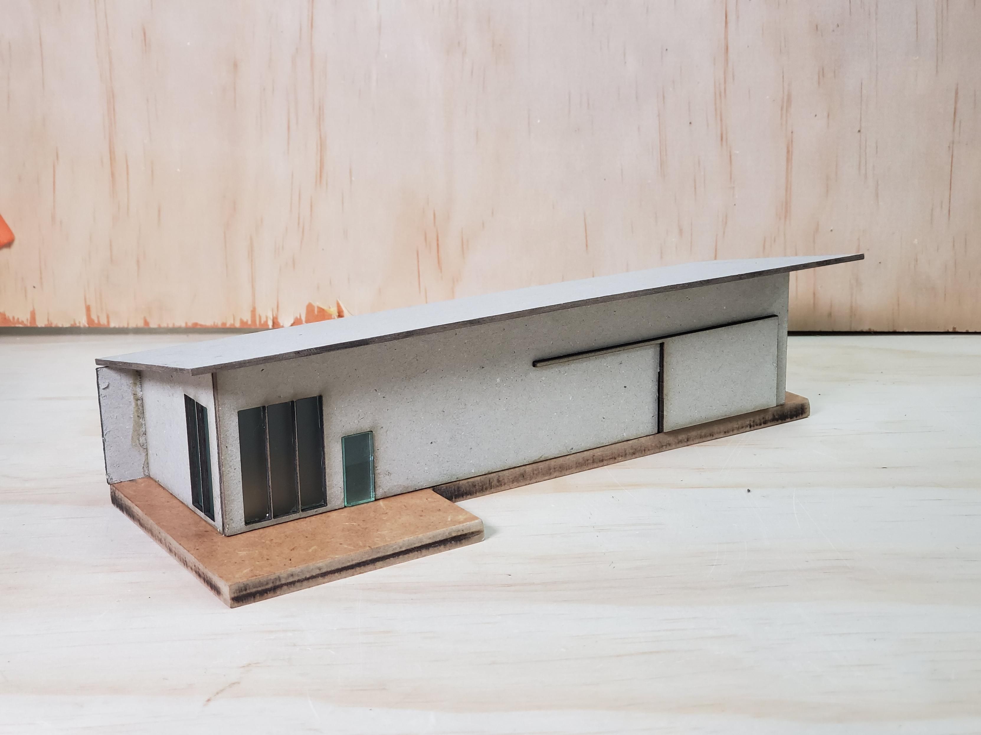 Chipboard model of a slant-roof style building with tall windows, a concrete patio, and a sliding door. 