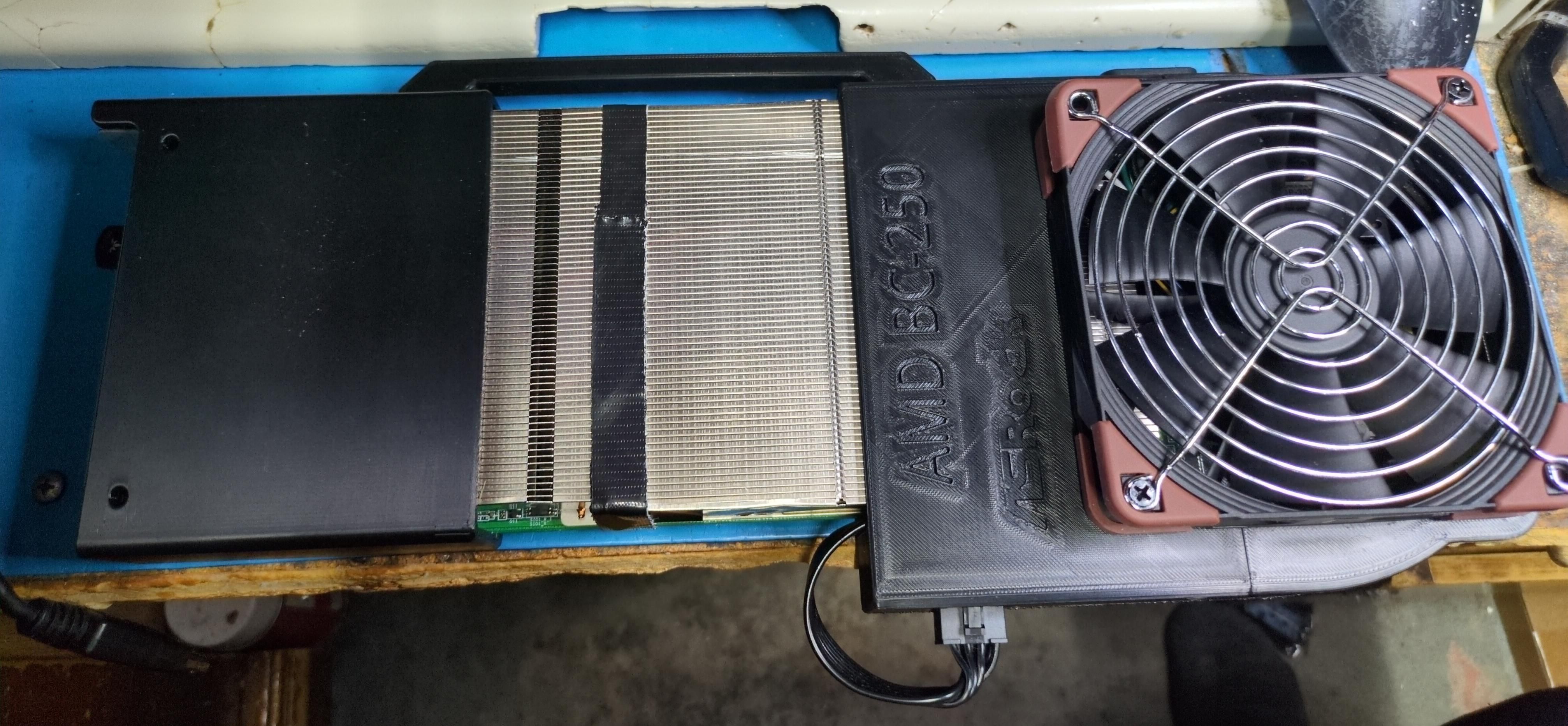 Picture of a device, shaped like a cube and roughly the size of a computer keyboard. There are 3D-printed shells on either end, with an exposed closed heat sink in the middle. A fan is attached to one of the shells. "ASROCK AMD BC-250" is engraved on the shell, as well.