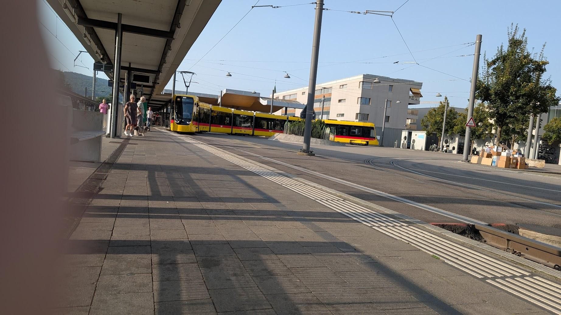 Photo of a yellow BLT tram, turning back towards Basel at Arlesheim/Dornach this morning.The sky is blue, there are buildings behind the tram.