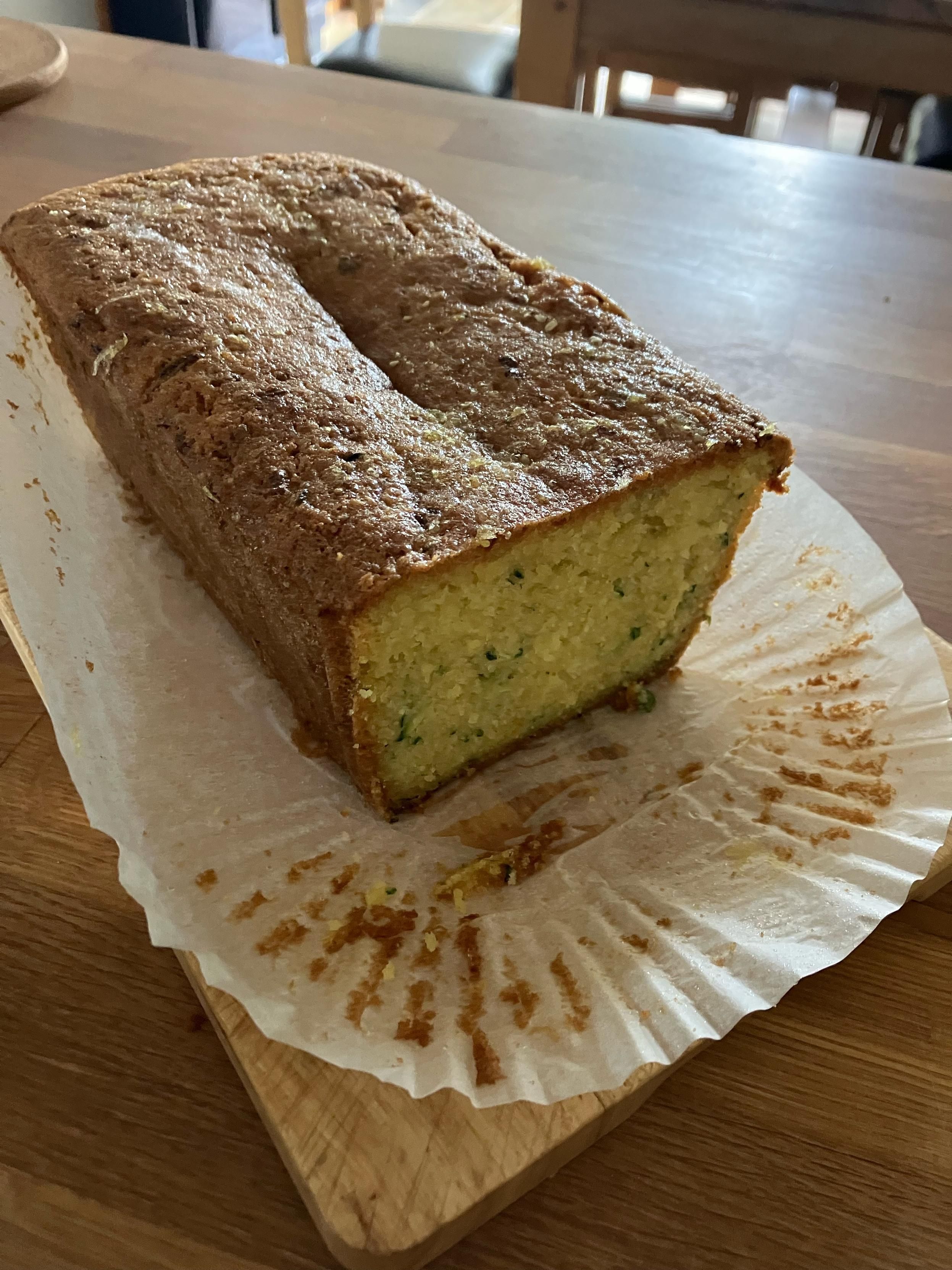 A photo of a loaf cake with the end sliced off. It’s golden brown on the outside, with a yellow sponge interior flecked with green. Courgette, thyme and lemon drizzle cake.