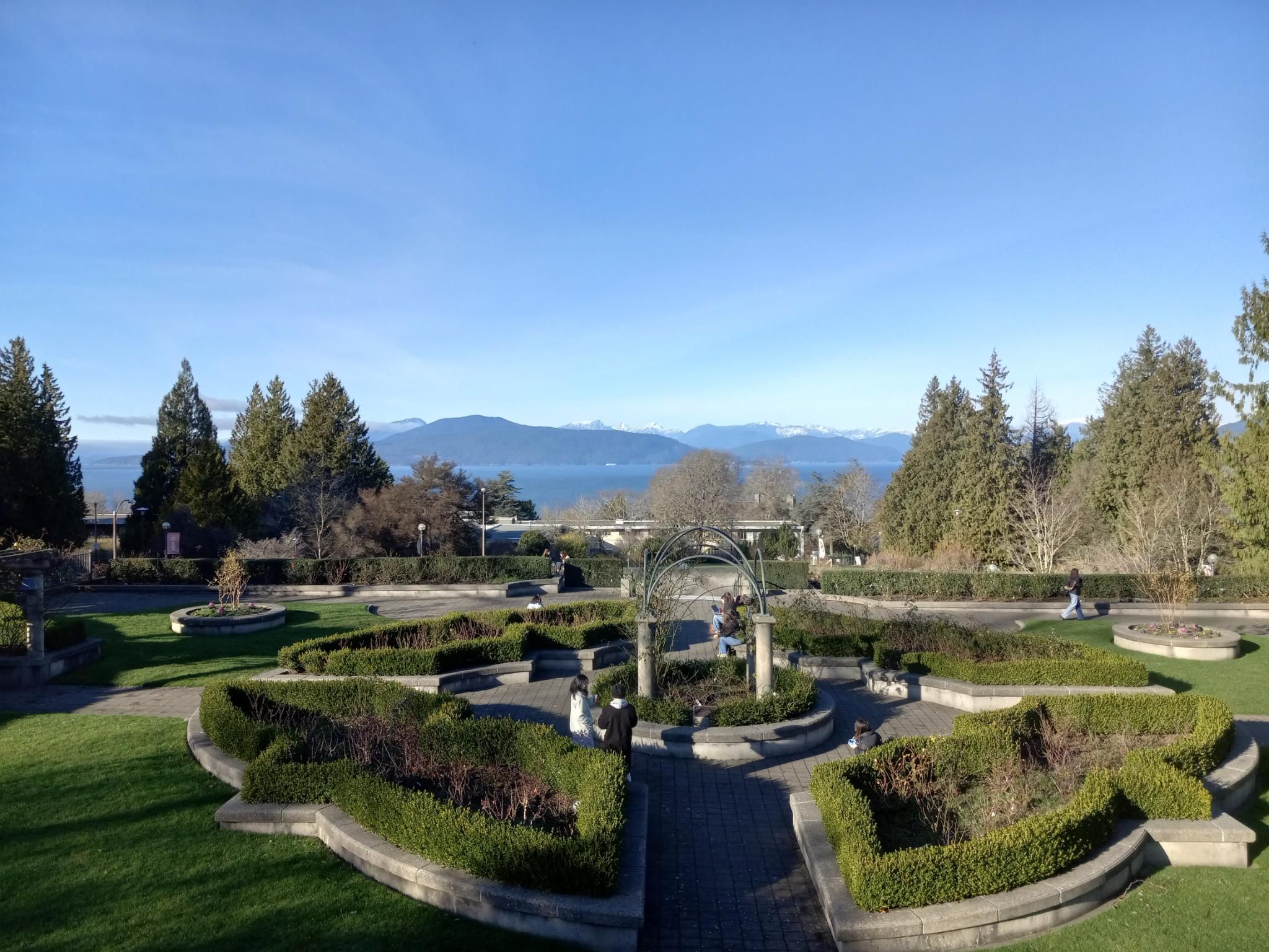 A ridiculously gorgeous view over a garden with shaped shrubs toward the bright blue ocean and distant snow-covered mountains, with the bright blue sky above.