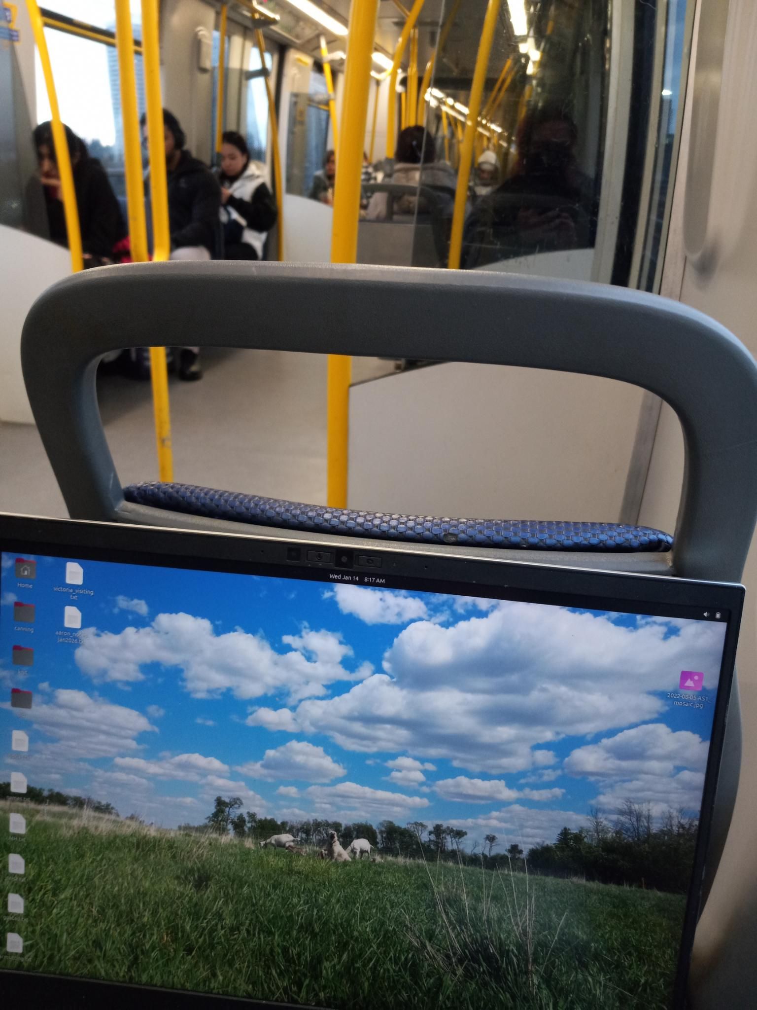 A view of my laptop on a fairly uncrowded skytrain.  My laptop has a pretty blue sky photo of a green grassy field on the desktop.  A few people are sitting in the distance down the traincar, and there are lots of yellow poles for standing train riders to grab onto.  