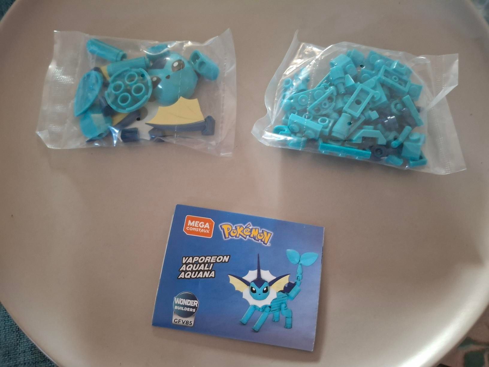 A picture of two little bags containing various blue pieces of a construction block toy. below the bags is a small paper pamphlet showing a completed Vaporeon in the Mega Construx line.