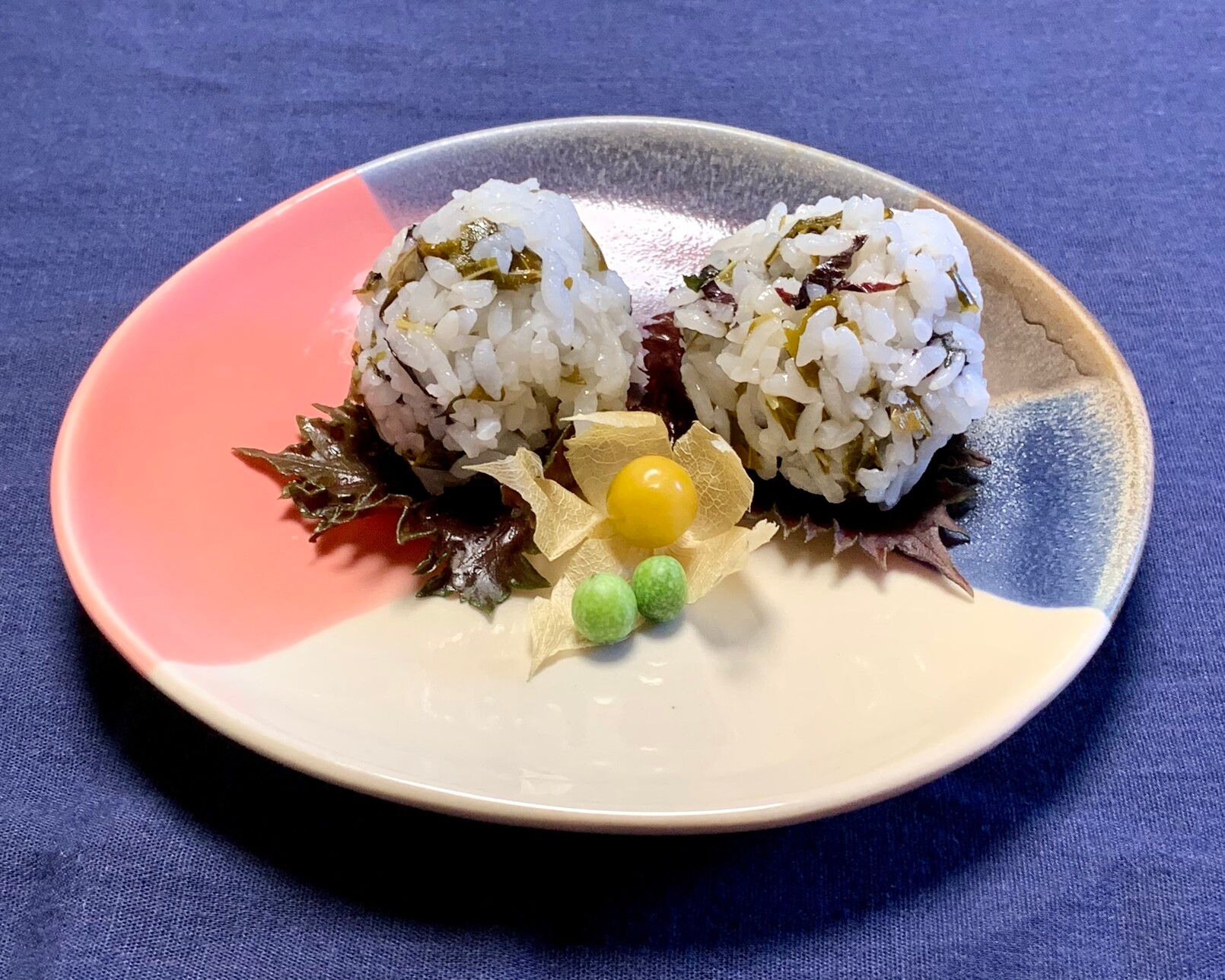 On a blue cloth is a irregular circular shape that is pie-graph colored from pink, navy, grey, lápiz and cream. On top of the plate are two onigiri balls that are rolled with sweet pickled strips of young jackfruit leaves with a little bit of shiso. They are on red and green shiso leaves. In front of them is a wild physallis opened from its paper casing like a yellow pearl and two tiny round green wild cucumbers. 