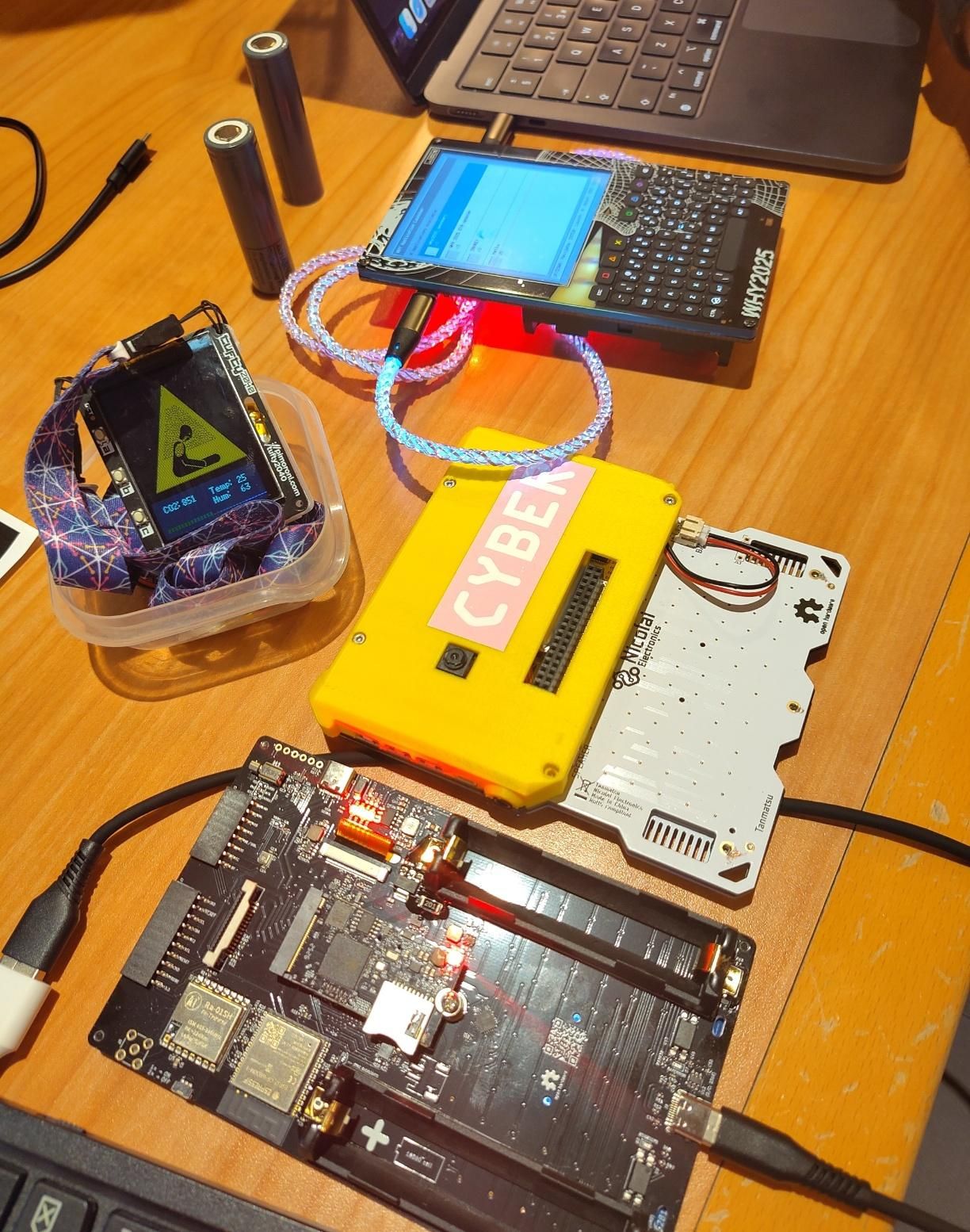 Photo showing two WHY badges and a Tanmatsu / Konsool, along with a DIY CO2 monitor badge. Some of the famously spicy WHY batteries and a couple of laptops can also be seen.