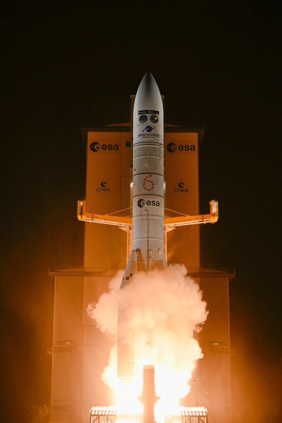 A Vega-C rocket launches at night from the European Space Agency’s facility, with bright flames and smoke billowing from its base. The rocket is marked with ESA, CNES, and Arianespace logos.