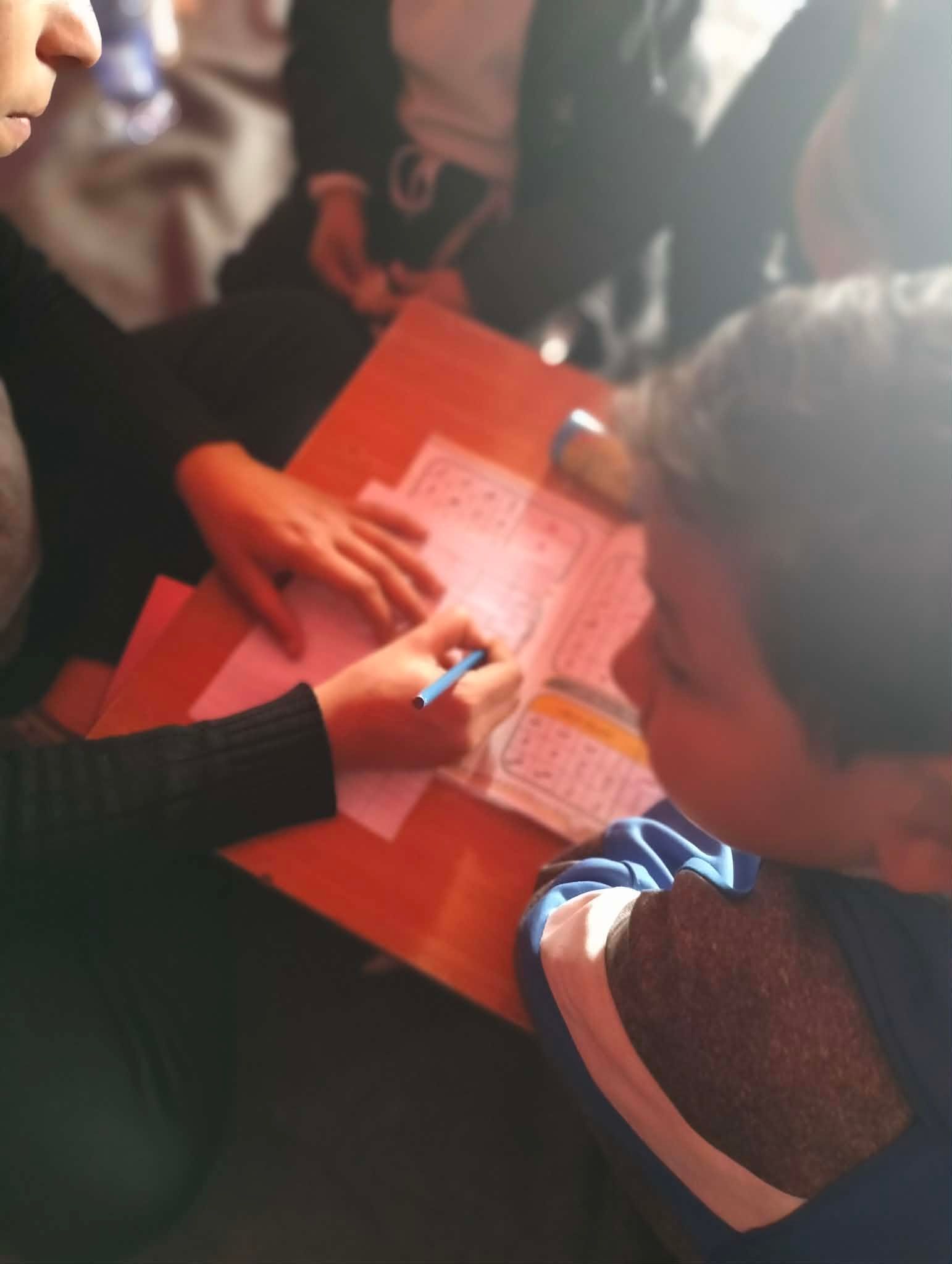 A person, writing with a blue pen on a sheet of paper with columns of printed numbers and symbols, on a wooden desk with other people. Another person is looking at the paper and person writing.