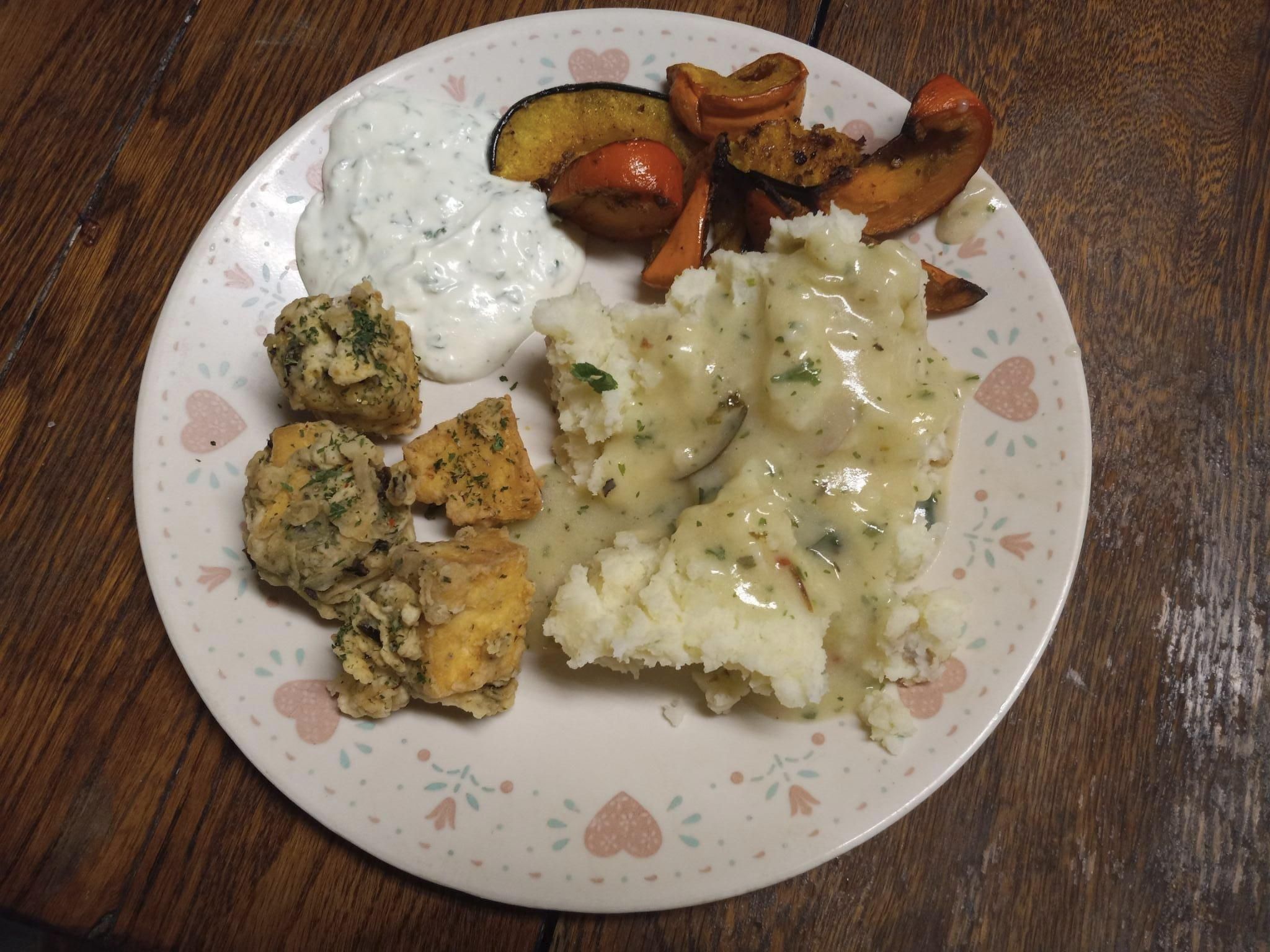 A view looking down at a plate full of food: fried tofu, mashed potatoes and gravy, baked squash slices, and garlicky yogurt sauce.  The plate has little hearts on it and its sitting on a wooden table