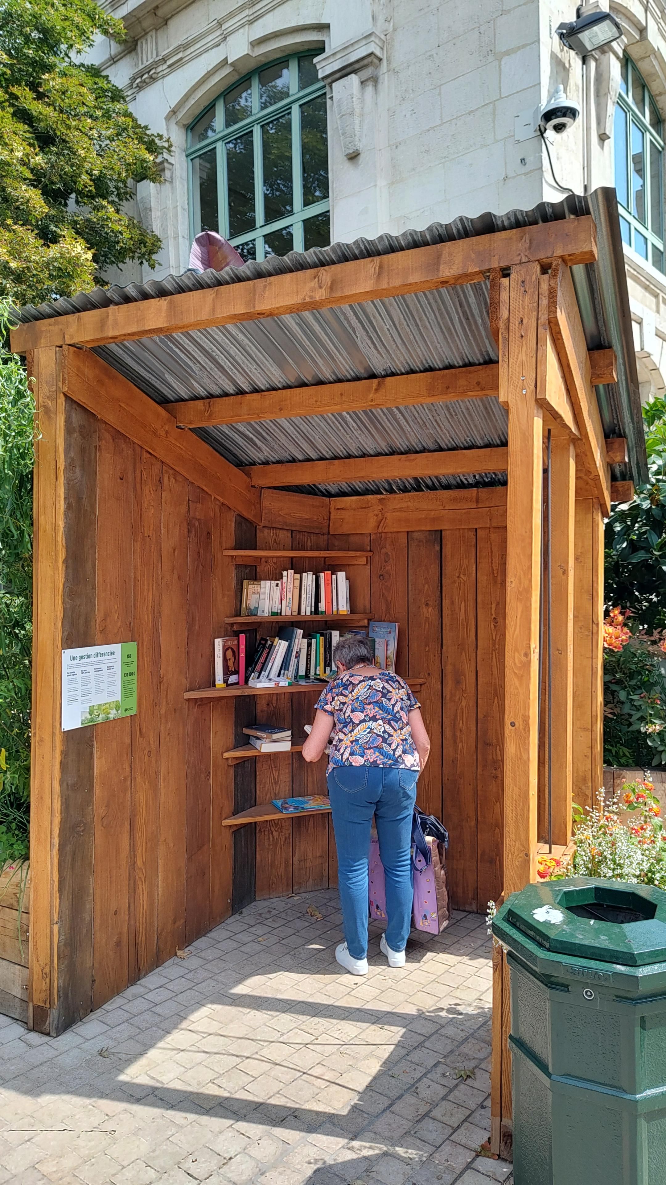 Hut van luxe houten balken in een stedelijke omgeving. In een hoekje zijn schuin een paar planken aangebracht, waarop boeken staan. Een oudere dame buigt zich over de collectie.