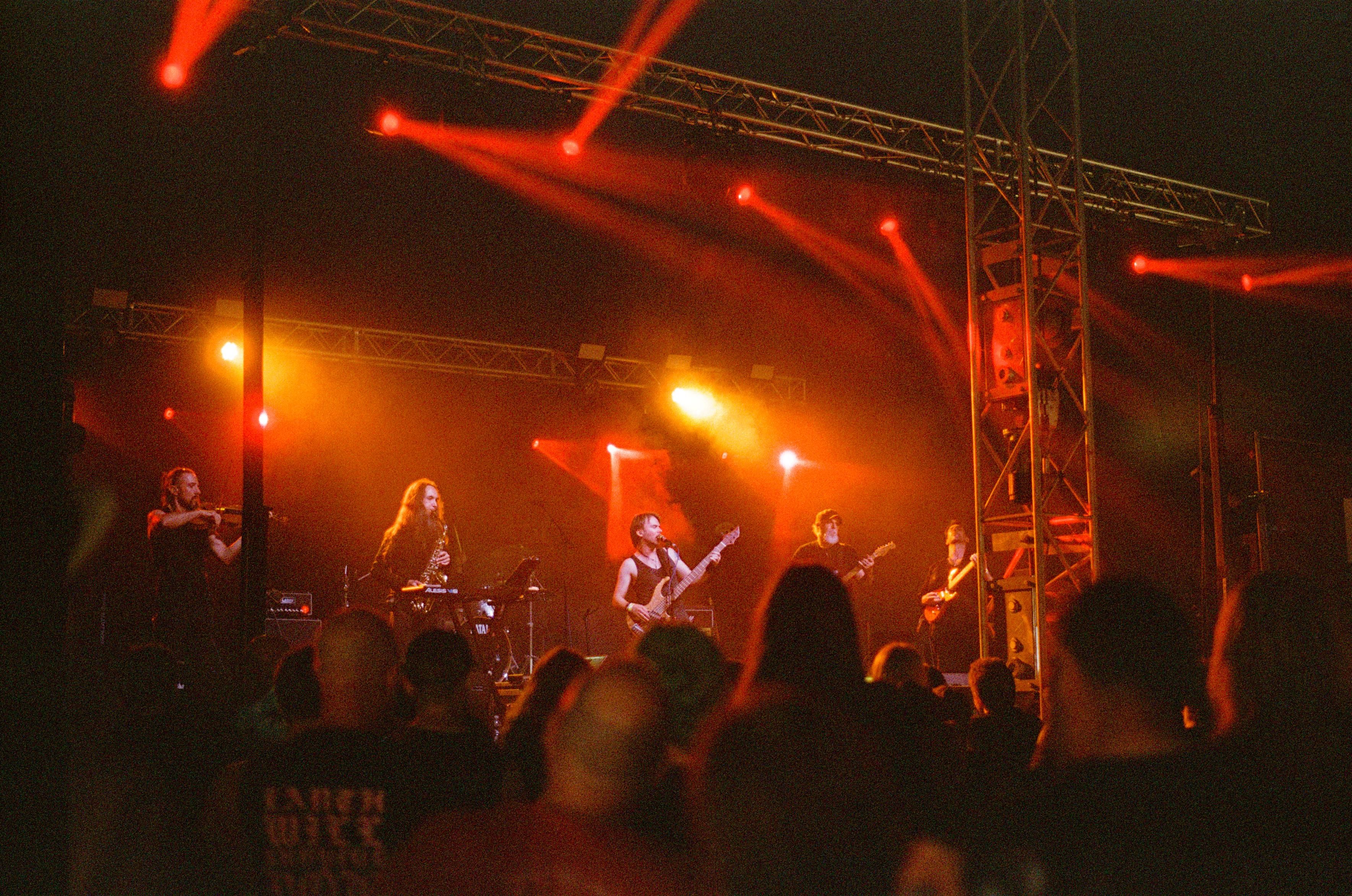 A band on stage illuminated by red lights. One person is holding a violin, another a saxophone, three more are holding guitars, the one in the middle is singing into a microphone. A crowd is visible in front of the stage.