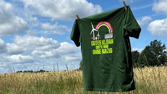 Ein T-Shirt auf einer Wäscheleine. Im Hintergrund Blauer Himmel mit weißen Wolkrn, Bäume und ein Feld mit Ähren. Das T-Shirt ist grün, auf ihm sind Regenbogen und Windräder zu sehen und die Aufschrift: "Gutes Klime gibt's nur ohne Nazis"