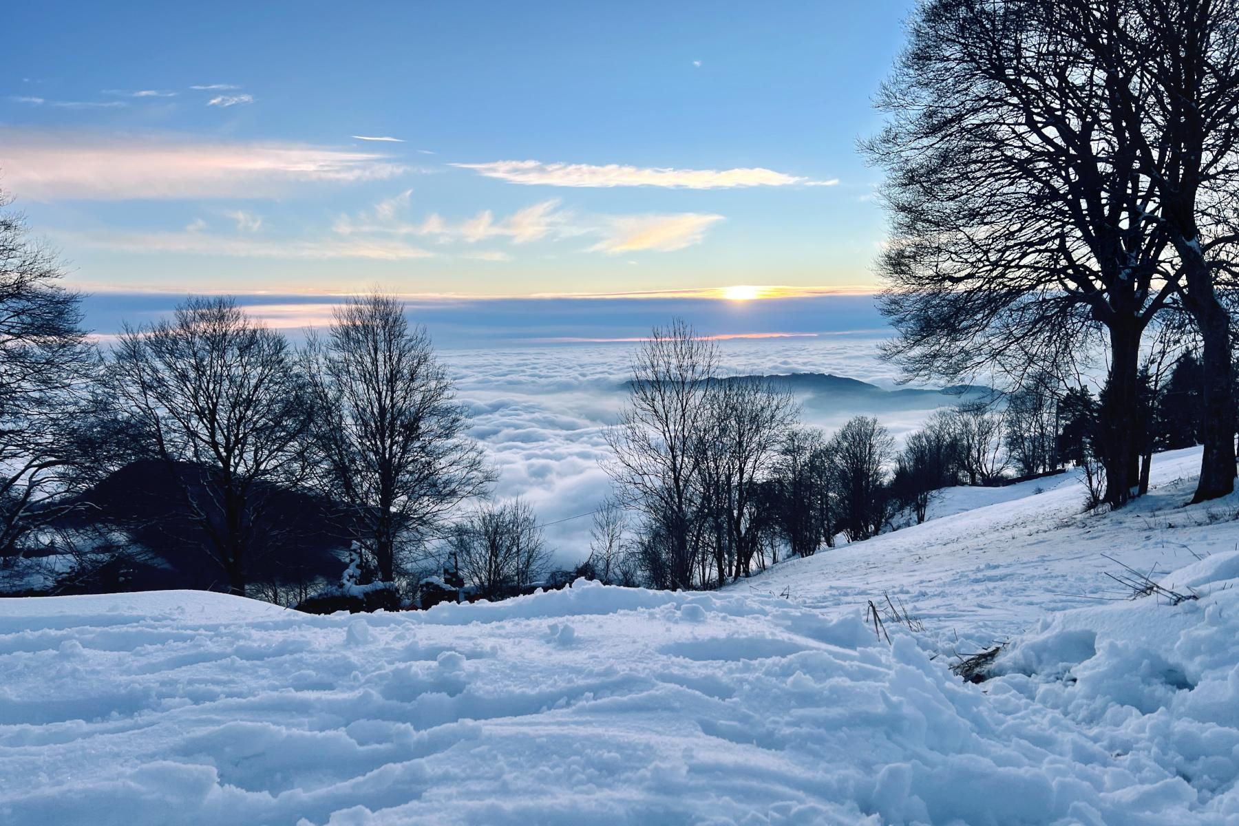 Paesaggio innevato al tramonto. Dall'alto della montagna si vede la valle sottostante ricoperta da nubi basse. 