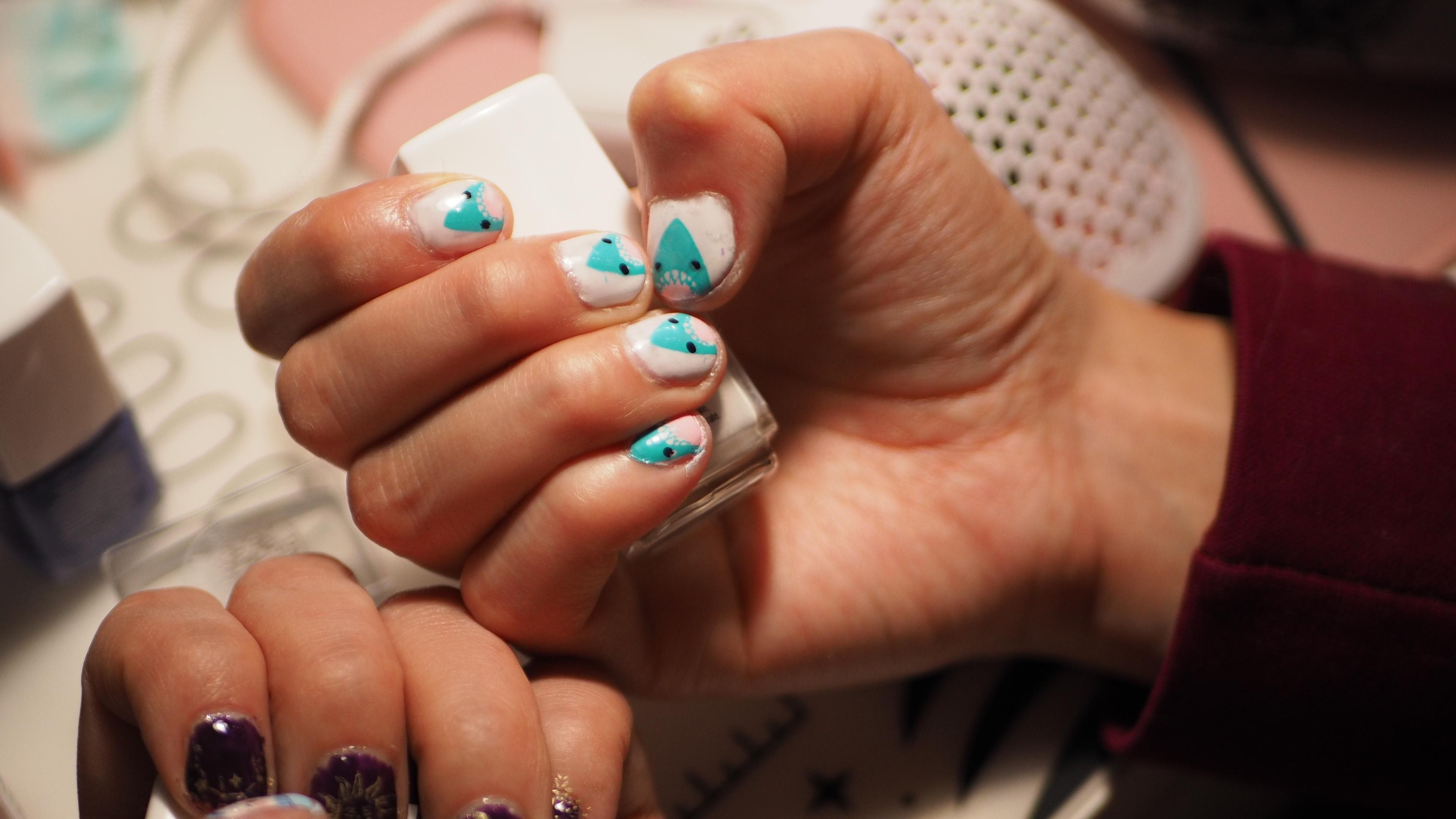 A hand holding a nail polish bottle, the nails are visible and are decorated with a blue shark with a pink belly and a very goofy look.