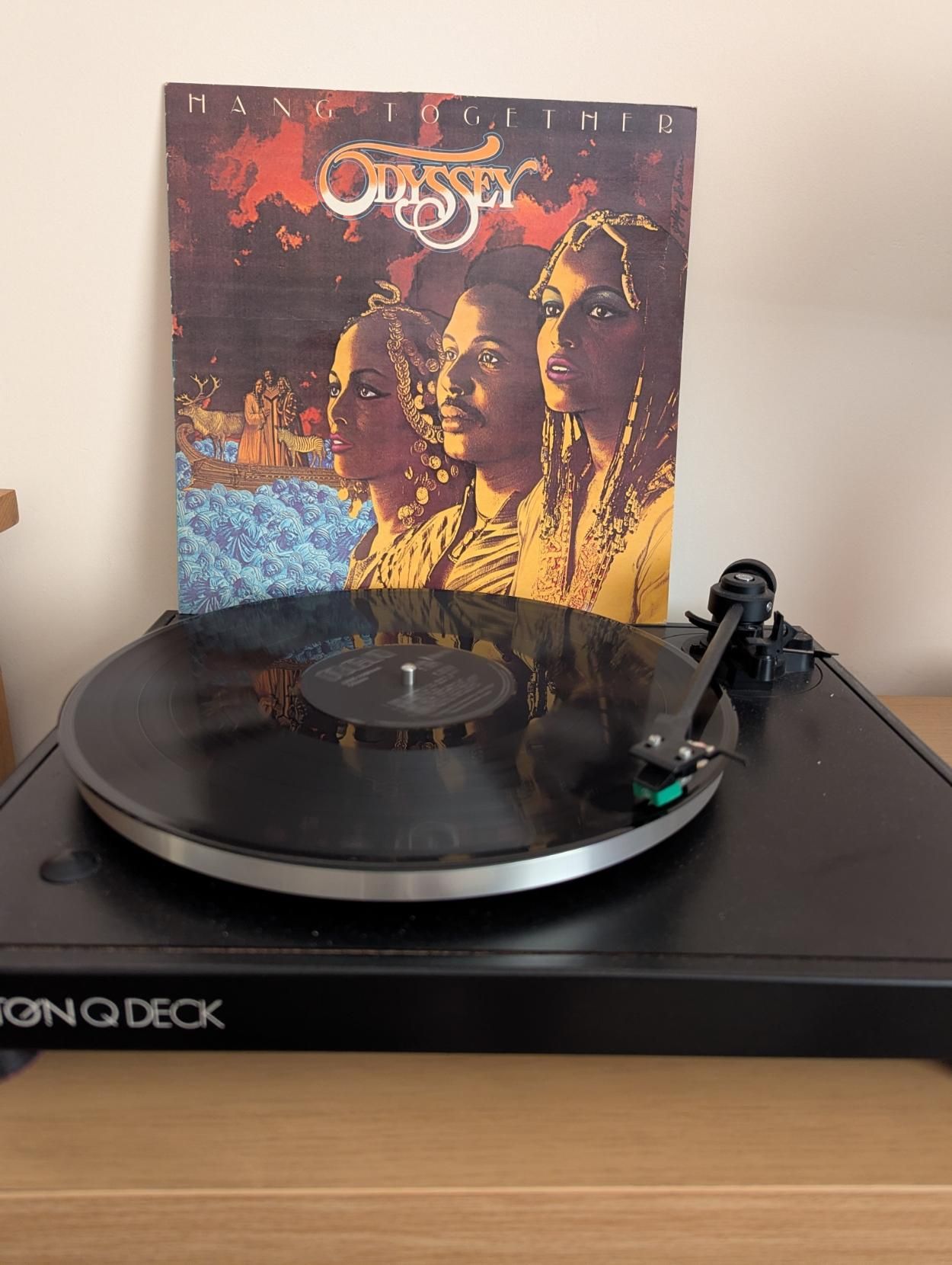A black vinyl record playing on the turntable with the album cover from Odyssey in the background featuring a drawing of the three main members