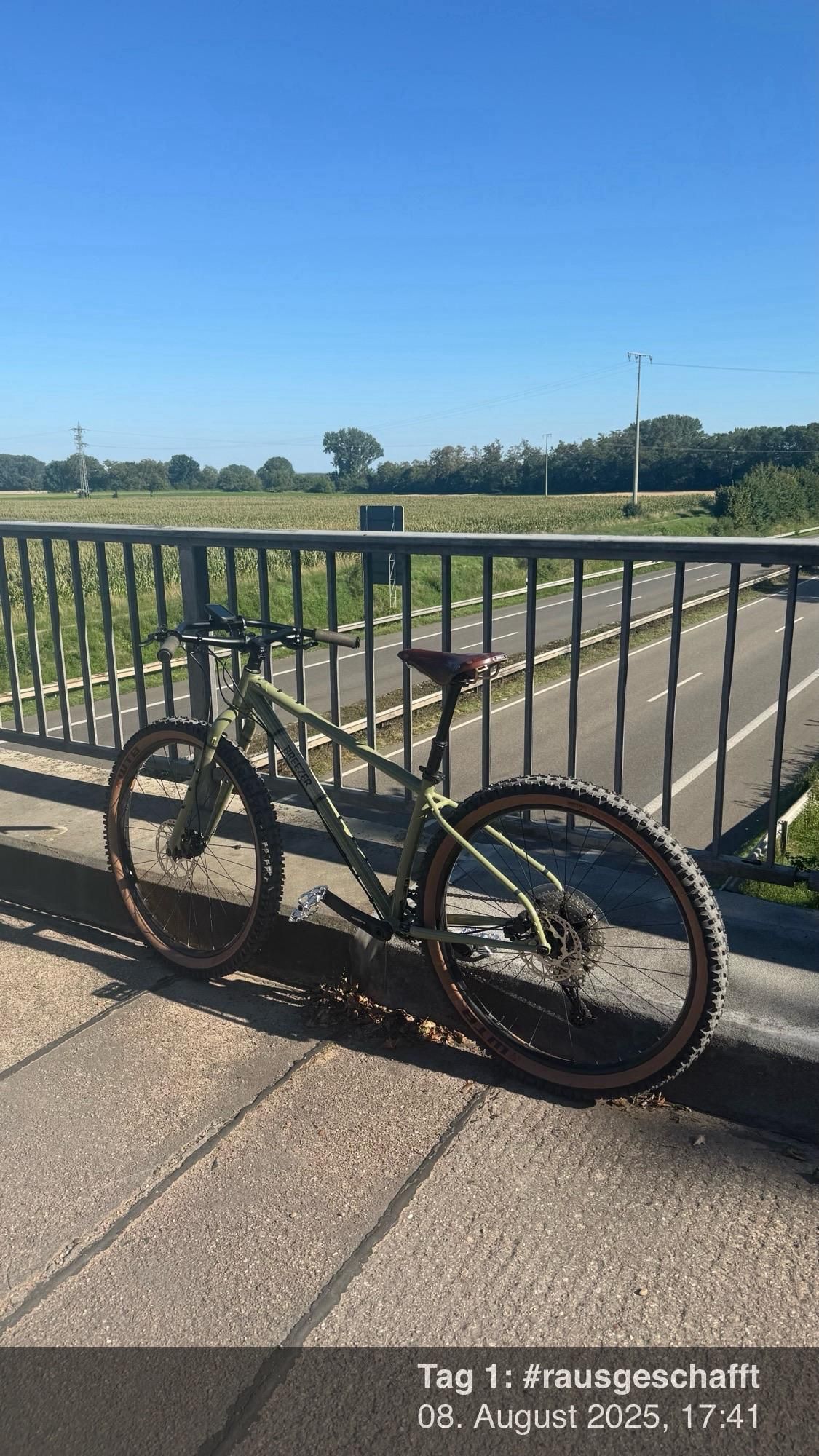 Ein grünes Mountainbike ist vor einem Geländer auf einer Brücke geparkt, mit Blick auf eine Straße und Felder unter einem klaren blauen Himmel. Die Umgebung weist auf eine ruhige Außenumgebung hin.