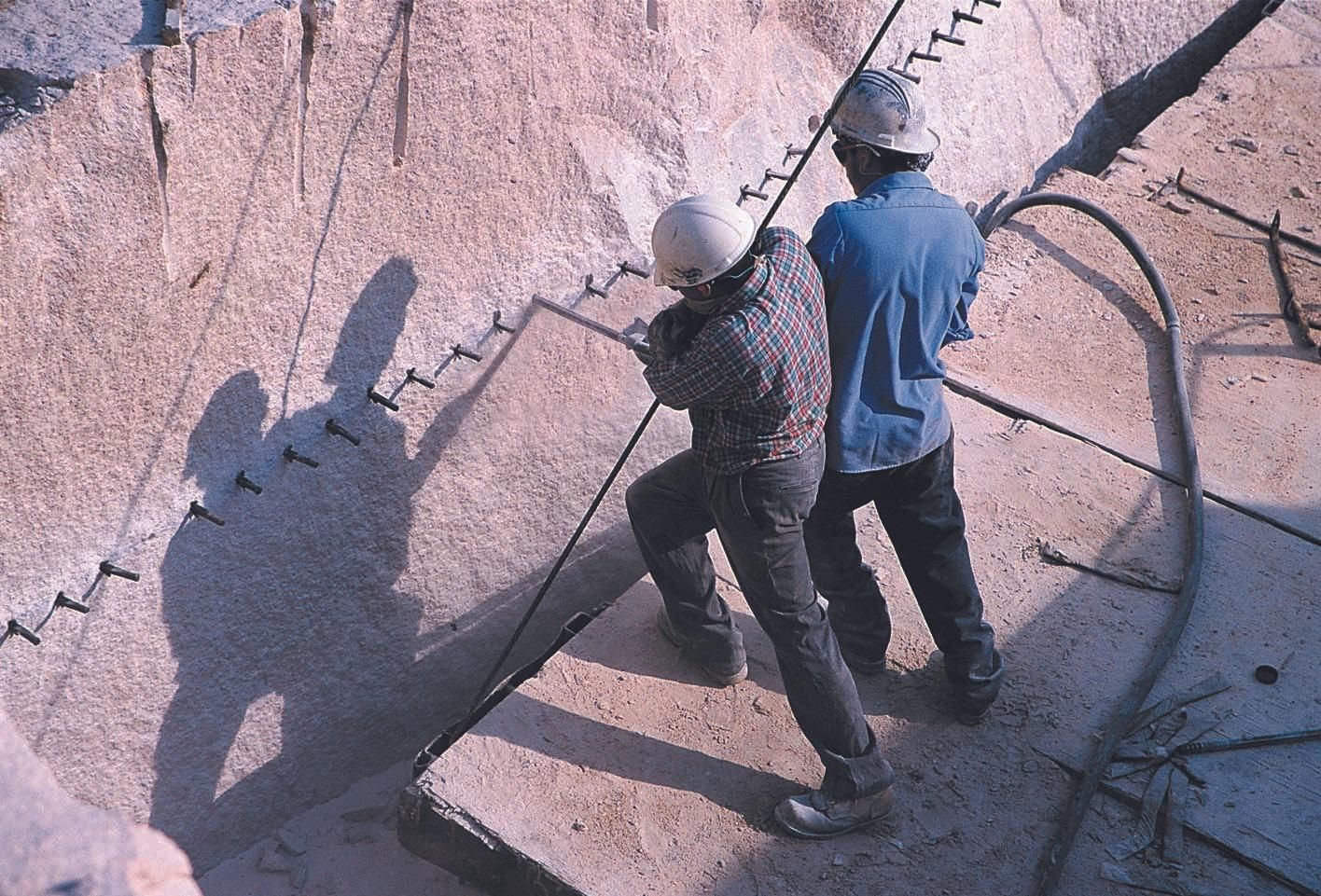Due operai perforano una superficie verticale, probabilmente per una volata di esplosivo.
La pietra sembra granito rosa.