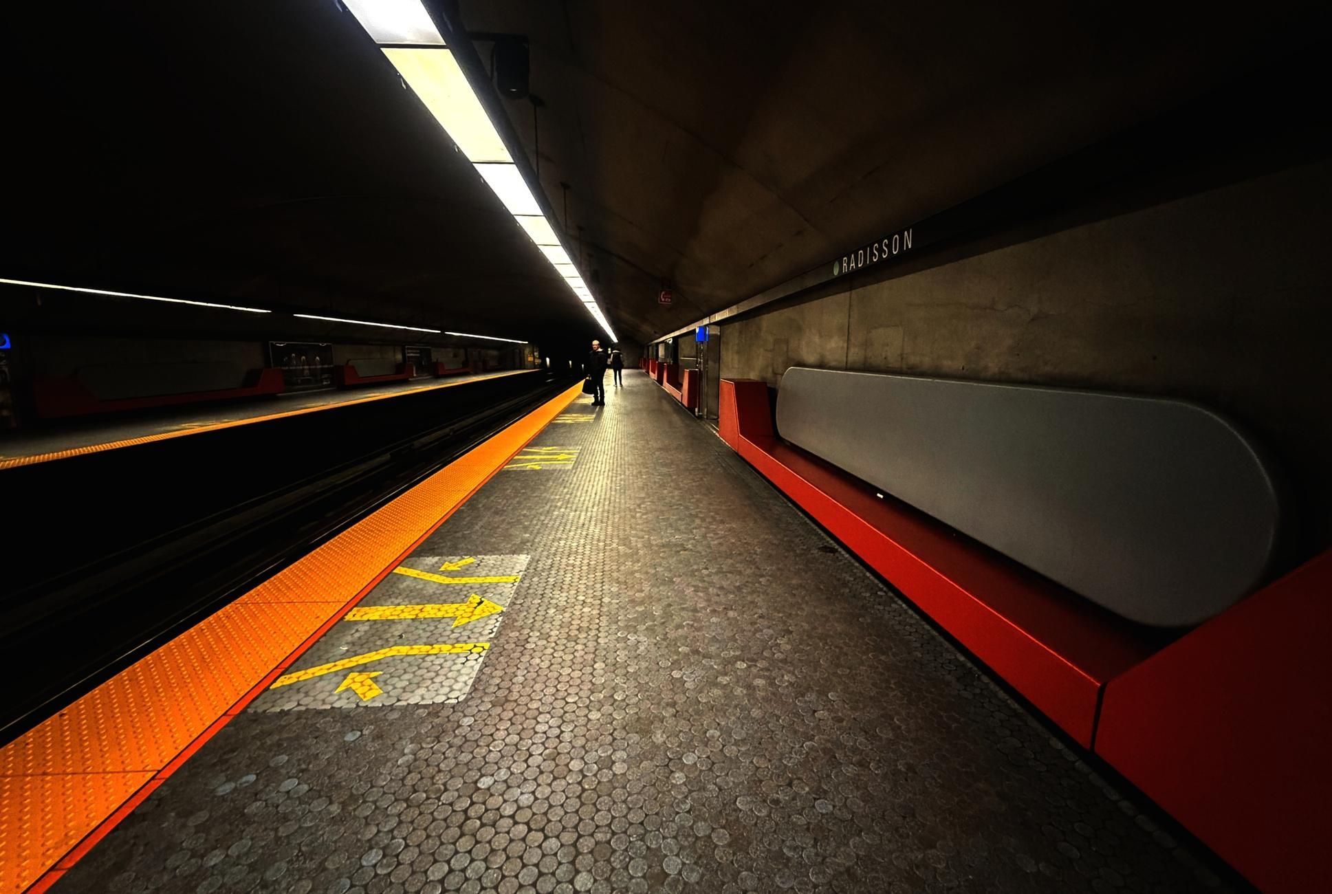 Photograph of an underground metro station, taken from one end of the platform looking towards the tunnel in the back, acting as a vanishing point. On the left, the tracks in the floor, the lights above and the reflective orange surfaces on the platform edges create lines extending into the background. In the center, the platform itself with its small, round blue tiles and yellow arrows indicating the train exit doors, and two passengers waiting at the far end. On the right, along the gray concrete wall, modern-style bench seating, with red seats and rounded gray backs, and the black band at the top with the station name "Radisson" in white, also create an alignment of shapes and colors towards the background.

Photographie d'une station de métro souterraine, prise à un bout du quai d'embarquement en regardant vers le tunnel au fond, qui agit comme point de fuite. À gauche les rails presque invisibles dans le sol, les lumières au-dessus des deux quais et les surfaces jaune-orange réfléchissantes sur le rebord créent des lignes allant vers le fond. Au centre le quai lui-même avec ses petites tuiles rondes bleues et les flèches jaunes indiquant les portes de sortie du train, et deux passagers en attente. À droite le long du mur de béton gris, des banquettes au design moderne, avec des bancs rouges et des dossiers gris arrondies, et la bande noire en haut avec le nom  "Radisson" en blanc, créent aussi un alignement des formes et couleurs vers le fond.