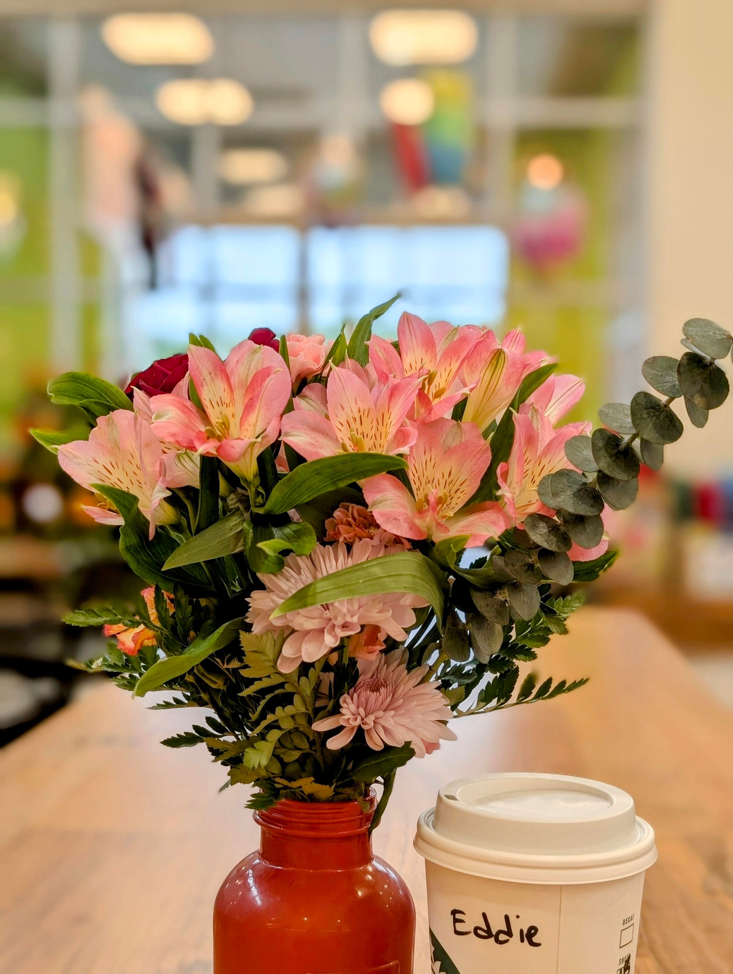 a small bottle urn with an arrangement of Peruvian lilies, mums, dried eucalyptus and fern sits on a long wooden table next to a carry-out coffee cup. The background is out of focus. store interior, large double-height windows to the outside and the rainy afternoon.