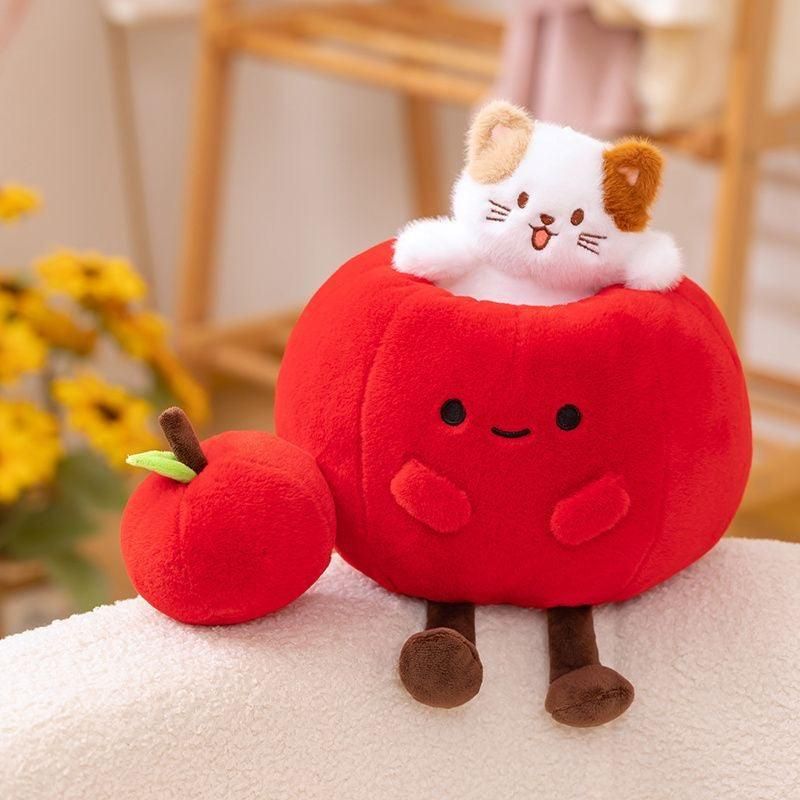 A plush white and brown calico cat popping out of a large, soft red apple. The apple has a simple smiling face and thin brown legs. A smaller, plain plush red apple sits next to it on a white textured surface.