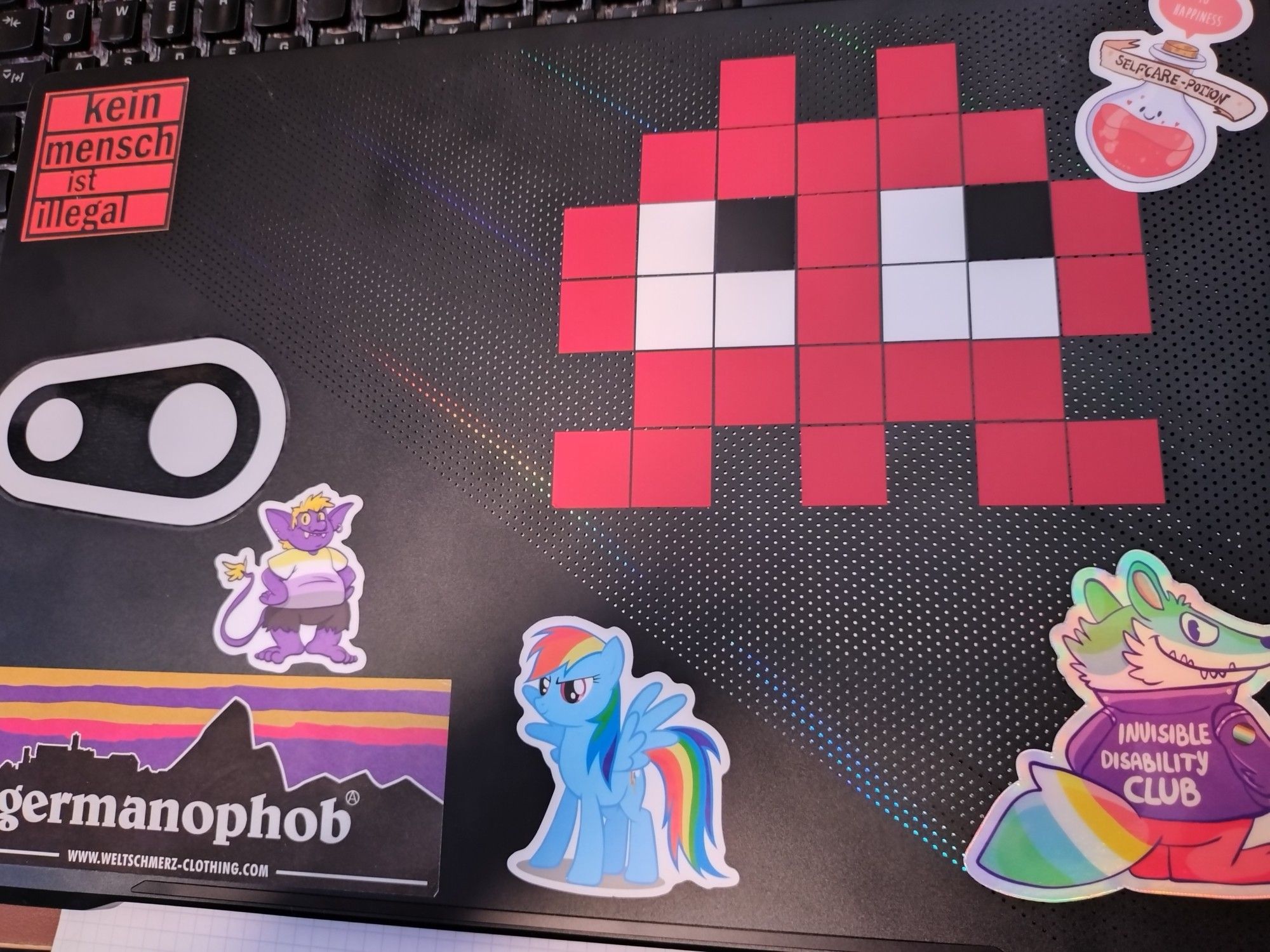 the back of my notebook display, again.
the 3 new stickers are a "Self-care potion + 10 happiness" a non-binary pride goblin and a holographic raccoon wearing an "Invisible Disability Club"-jacket

old stickers are a "No human is illegal", the crankbrothers logo, an "germanophob"-sticker looking like the Patagonia-logo, rainbow dash and a pixeled ghost from individual squares