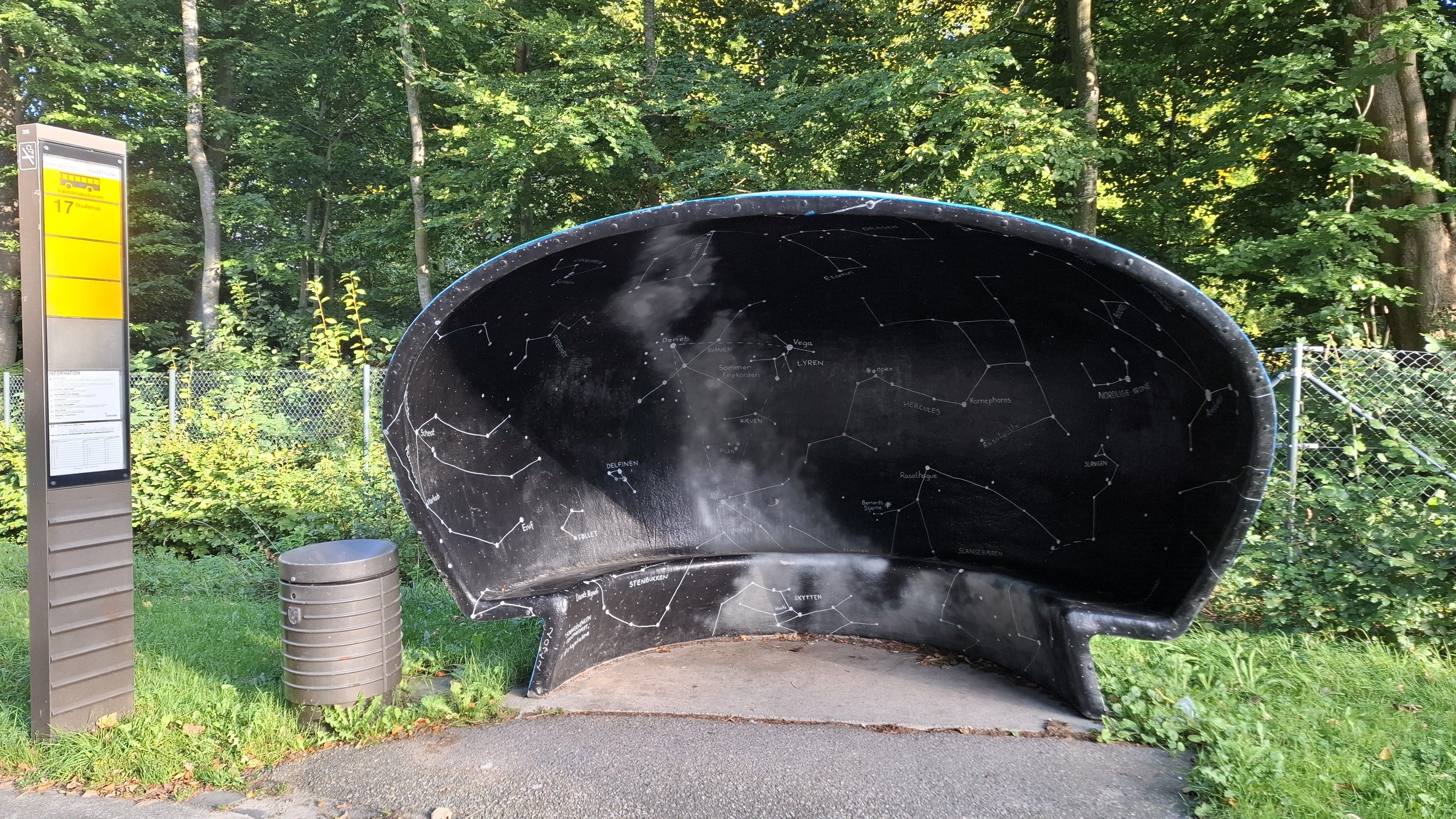 Aarhus. On the road outside the Ole Rømer Observatory, named after another famous Dane, Carl Nielsen. A broad clear road in Kongelunden, the Royal Woods, with trees on either side. Looking straight at the bus shelter, one of the city’s iconic mushroom-shaped bus shelters designed in the 1960s by furniture designer Poul Cadovius. A low concrete mushroom-shaped shell with standing room and a little bench inside. The council commissioned artists to paint them with local motifs.  Artist Ekaterina Poliakova painted the night sky in this one. As you wait for the bus, you are surrounded on three sides and above by a black night sky with a map of the stars and constellations and galaxies, their names written beside them.  Outside, the shelter’s shell is painted like a day sky – light blue with fluffy white clouds. 