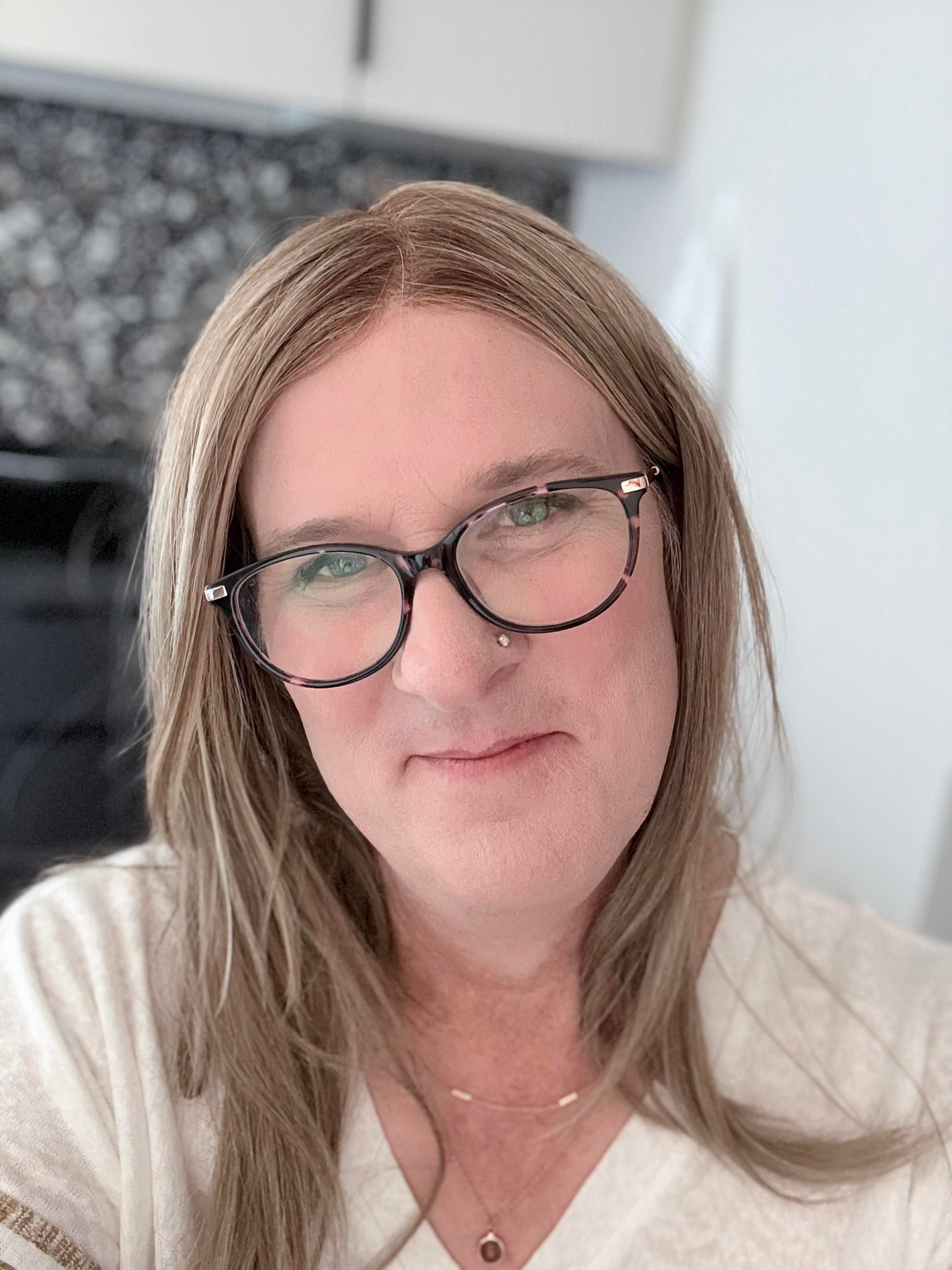Photo of a smiling Sophie wearing her usual black and pink cat eyed glasses. She has beyond shoulder length blonde hair, is wearing a cream colored v-neck top, and two gold necklaces. She is thinking after that last selfie, “Holy shit I’m a girl!”