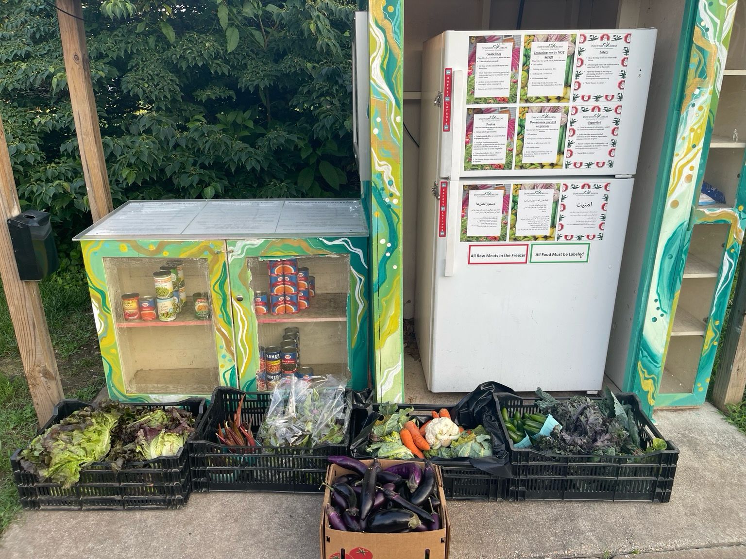 Four large crates and a box of various produce including leafy greens, brassicas, carrots, peppers, etc in front of a free fridge and community pantry. The fridge is closed and the pantry has about thirty canned goods in it from previous contributions.