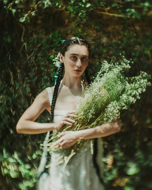 A feminine figure in a simple, loose white dress with long brown hair hanging in plaits over each shoulder, her arms  up cradling a posy of delicate white flowers. Her face is in focus as she looks a the camera, the outside of the image blurred with a hint of movement.