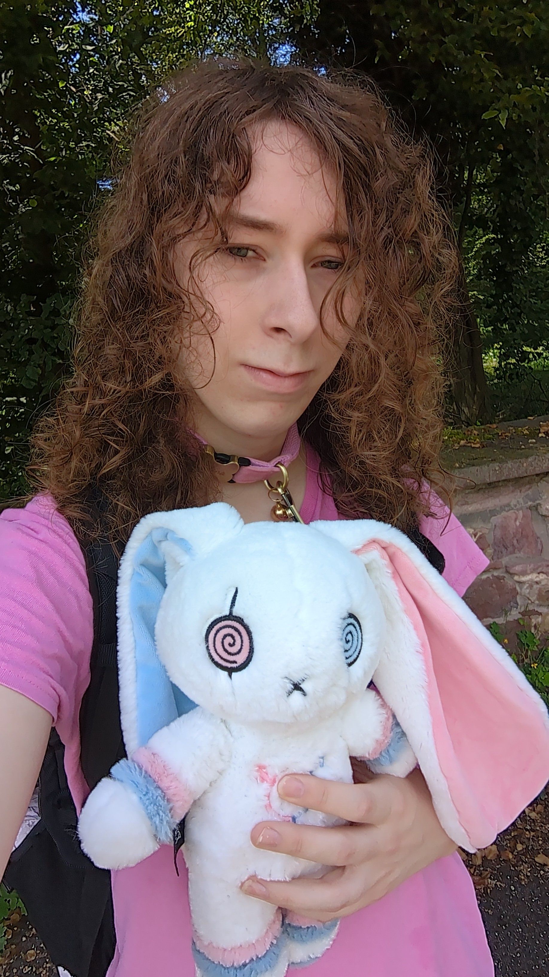 alina with darker, longer curly hair, wearing a pink collar and tshirt and her trans pride bunny in front of a forest