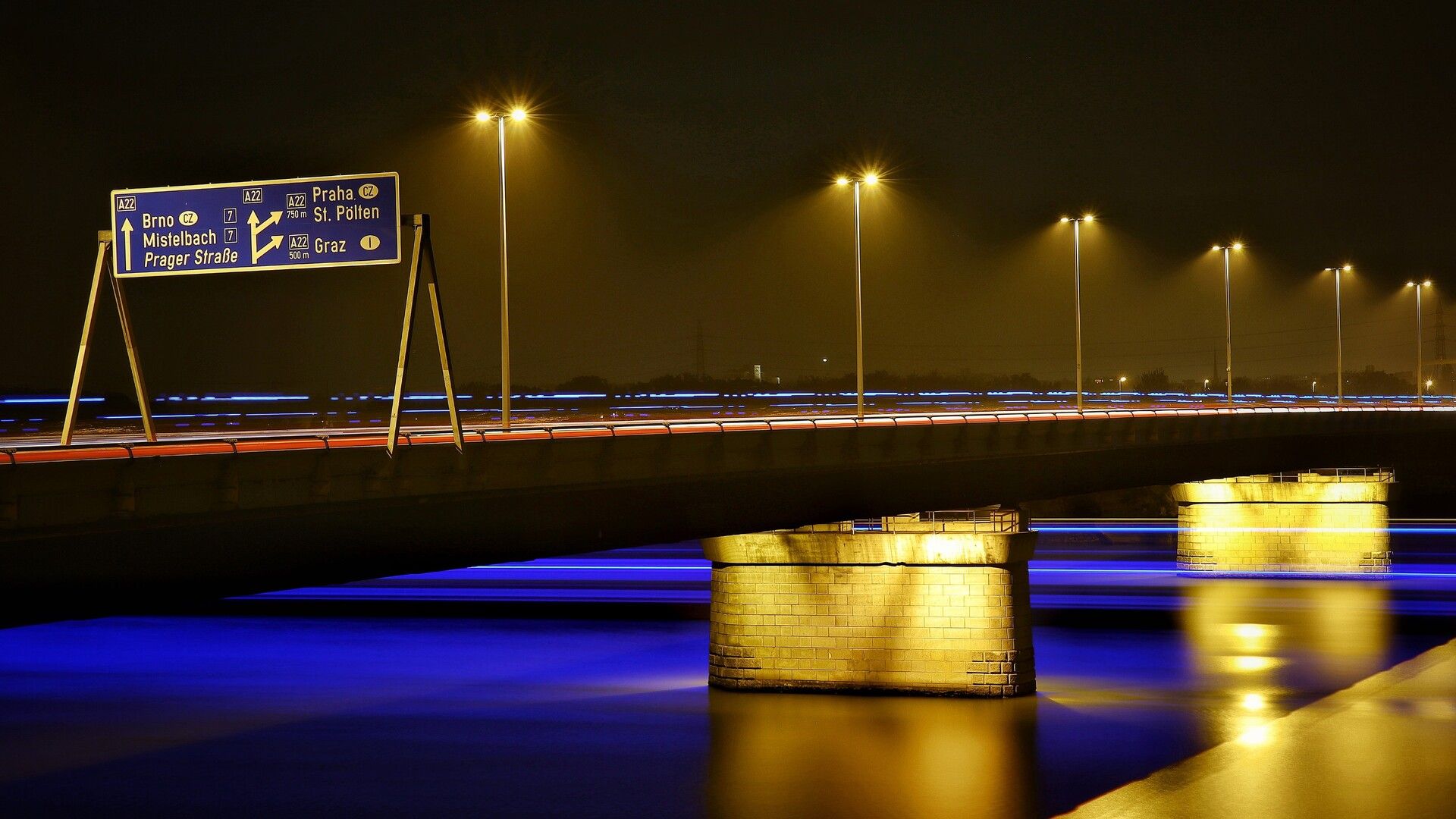 Es ist Nacht und man sieht eine Autobahnbrücke, die über eine Wasserfläche führt. Da es eine Langzeitbelichtung ist, ziehen auf und unter der Brücke diverse Fahrzeuge und Schiffe lange Lichtstreifen durch das Bild. Deutlich zu erkennen ist ein großer Überkopfwegweiser mit Zielen wie z.B. Brno, Mistelbach, Praha und St. Pölten. Die Lichter der Straßenbeleuchtung verschwinden leicht im Nebel.