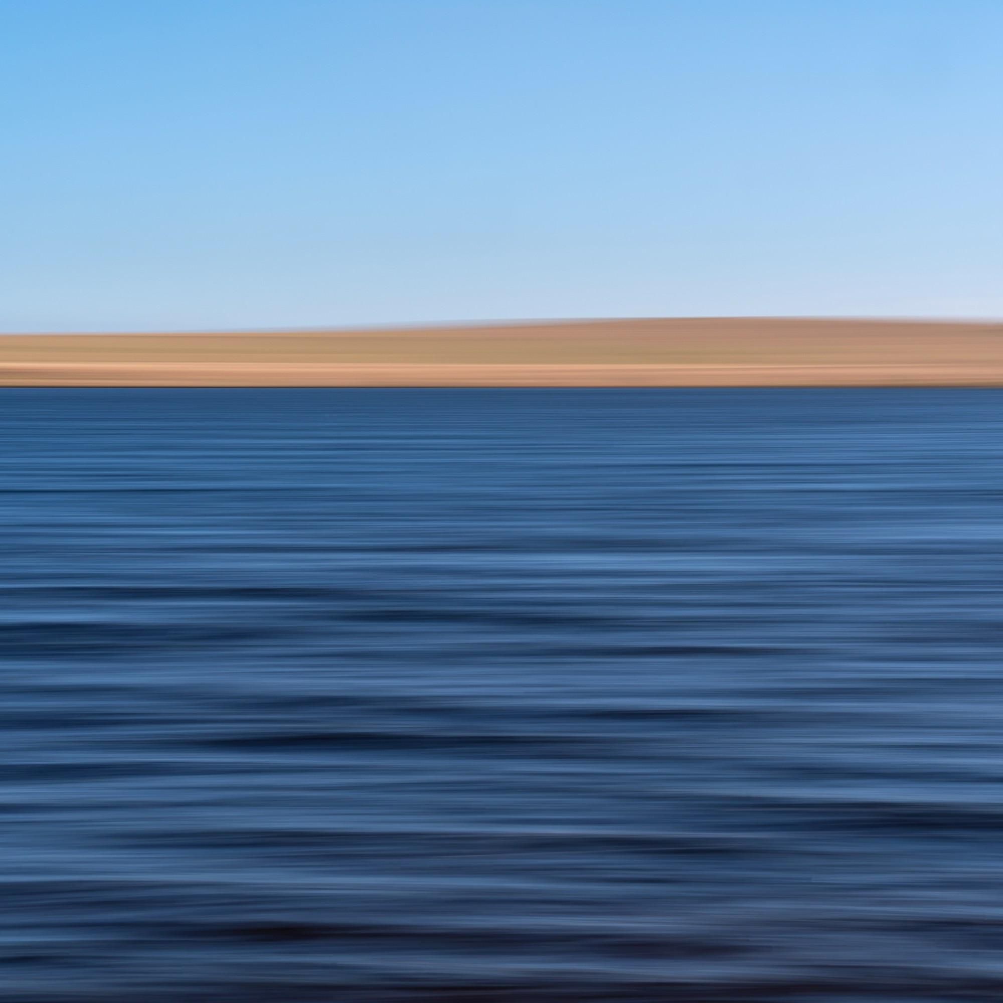 A tranquil view of deep-blue coloured water of a lake, stretching to meet a distant shore. A gentle rolling hill, in the light brown colour of dry summer grass under a clear blue sky, two-thirds up the frame. Horizontal ripple lines on the water create a sense of calm and expanse.