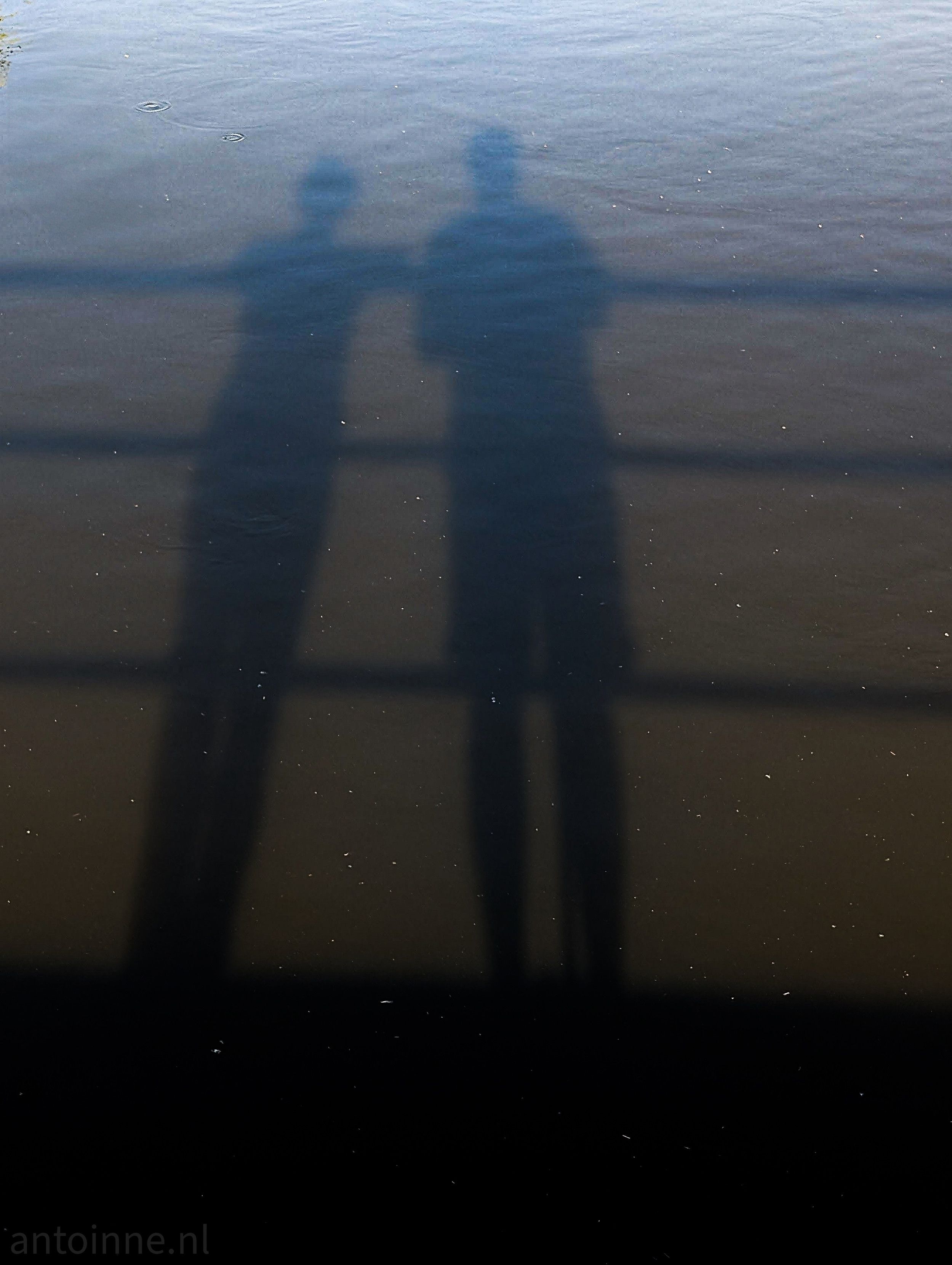
A photograph showing the shadows of two people cast onto a body of water. The shadows are long and dark, suggesting the sun is low in the sky. The two figures are standing next to each other, with the shadow on the left being slightly smaller and leaning on a railing. The shadow on the right is taller and stands upright.

The railing itself casts horizontal and vertical shadow lines across the water, creating a grid-like pattern. The water surface is dark and reflective, with some ripples visible, and a lighter, more blurred area at the top of the frame. There's a subtle reflection of the sky.
