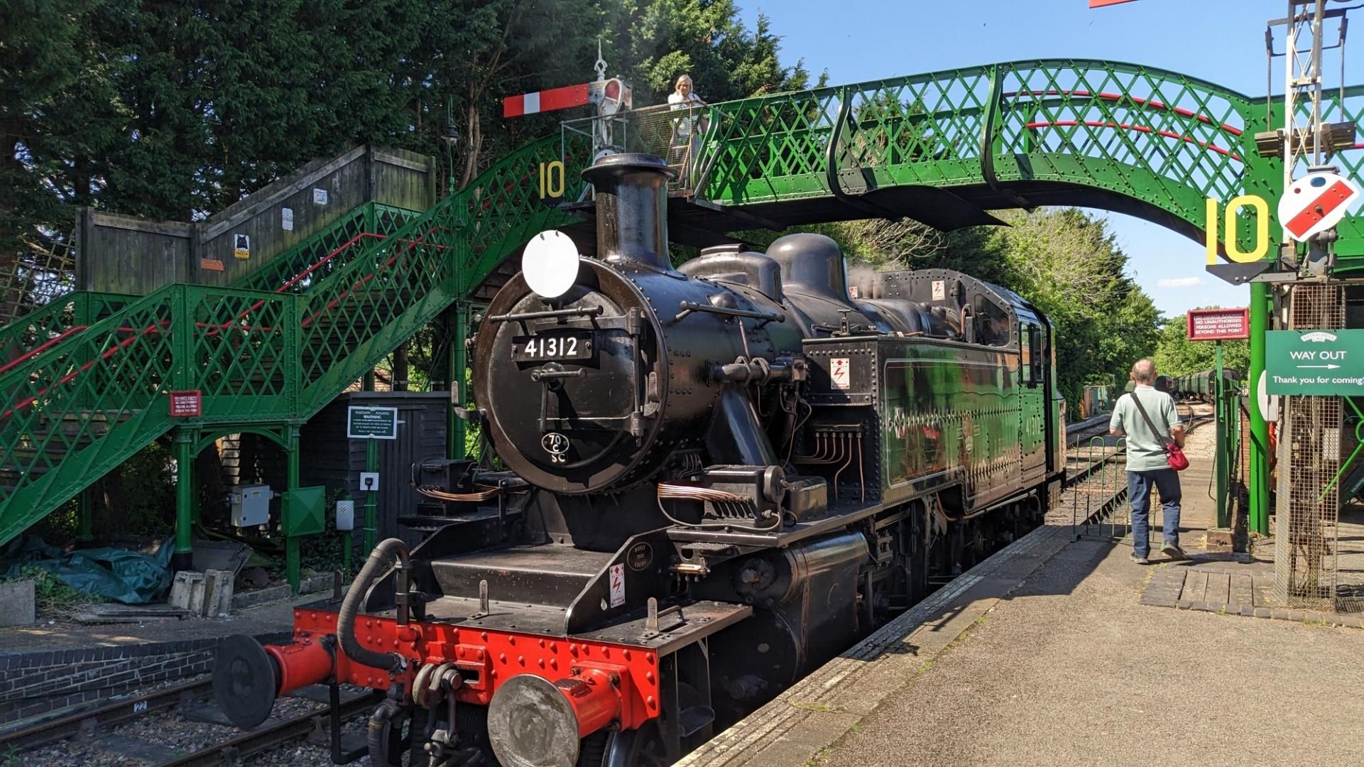Black steam loco pulling under a green steel footbridge with signals left and right