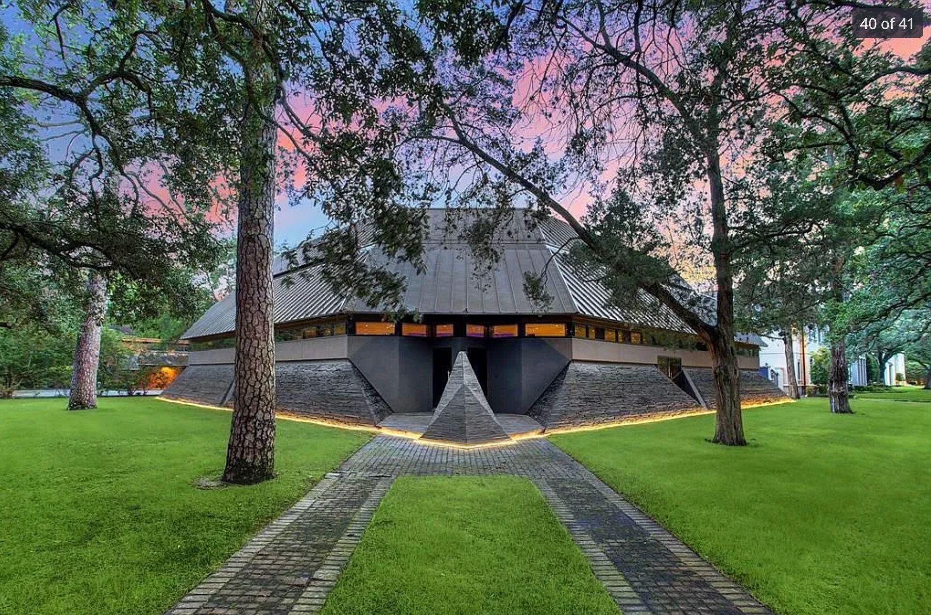 A house that looks like some sort of space ship with very small and narrow orange lit windows, 2 paths to the front door which has a concrete triangle in front of it, and the roof takes up most of the house. It is in dark brick and is ominous. 