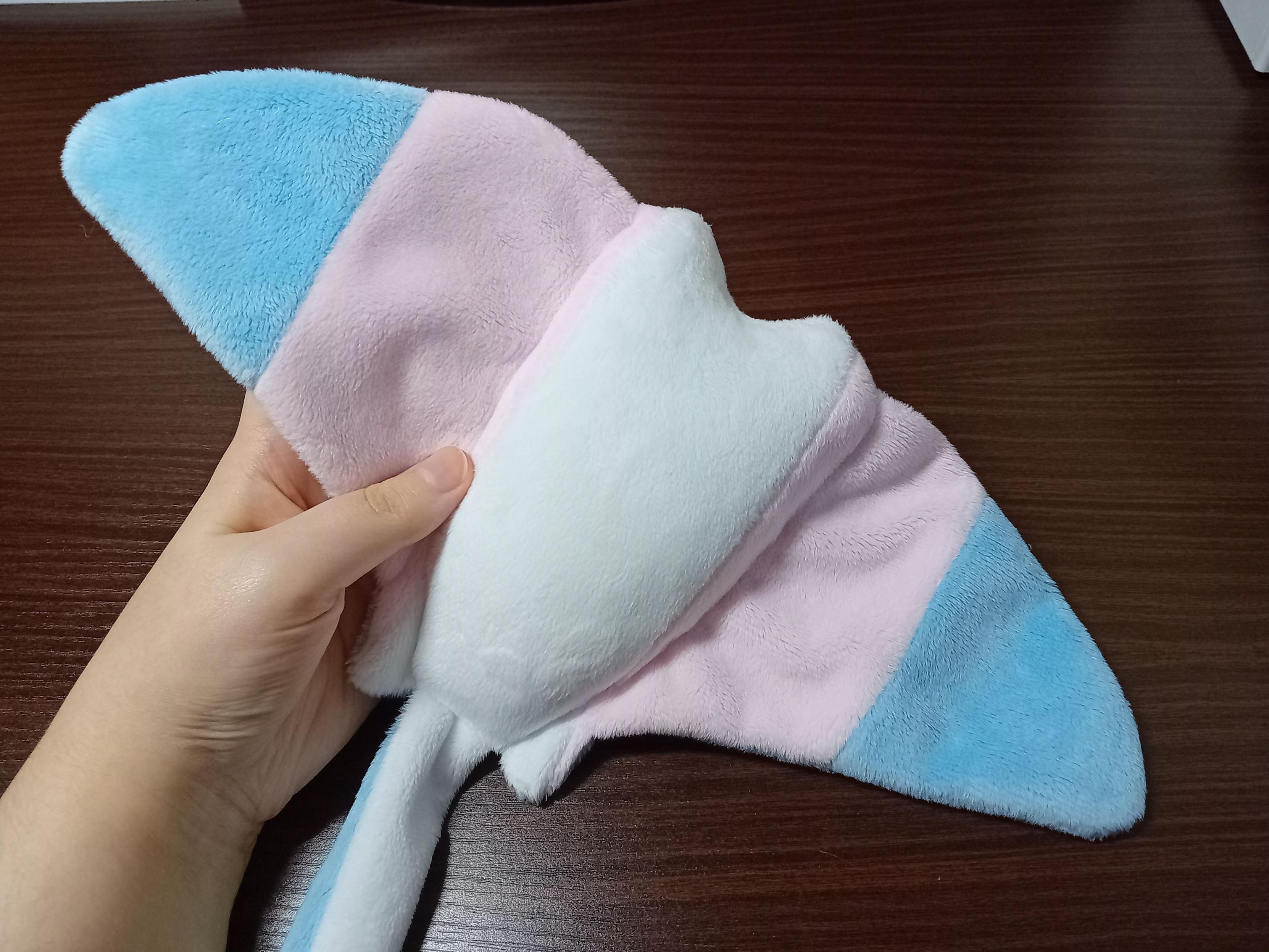 My hand holding a plushie trans flag Seaflap (stingray), it's flipped onto its belly so you can see the blue, pink and white stripes of the flag going from wing tip to wing tip.