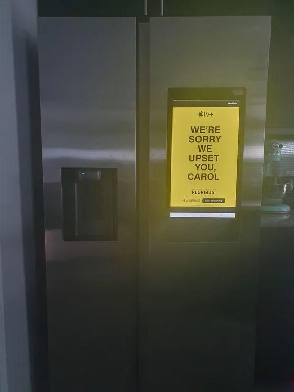 A stainless-steel, side-by-side refrigerator with a built-in touchscreen display on the right door. The screen shows a bright yellow Apple TV+ advertisement reading “WE’RE SORRY WE UPSET YOU, CAROL” for the series Pluribus. The left door includes a water and ice dispenser. The fridge is in a modern kitchen with small appliances visible in the background.