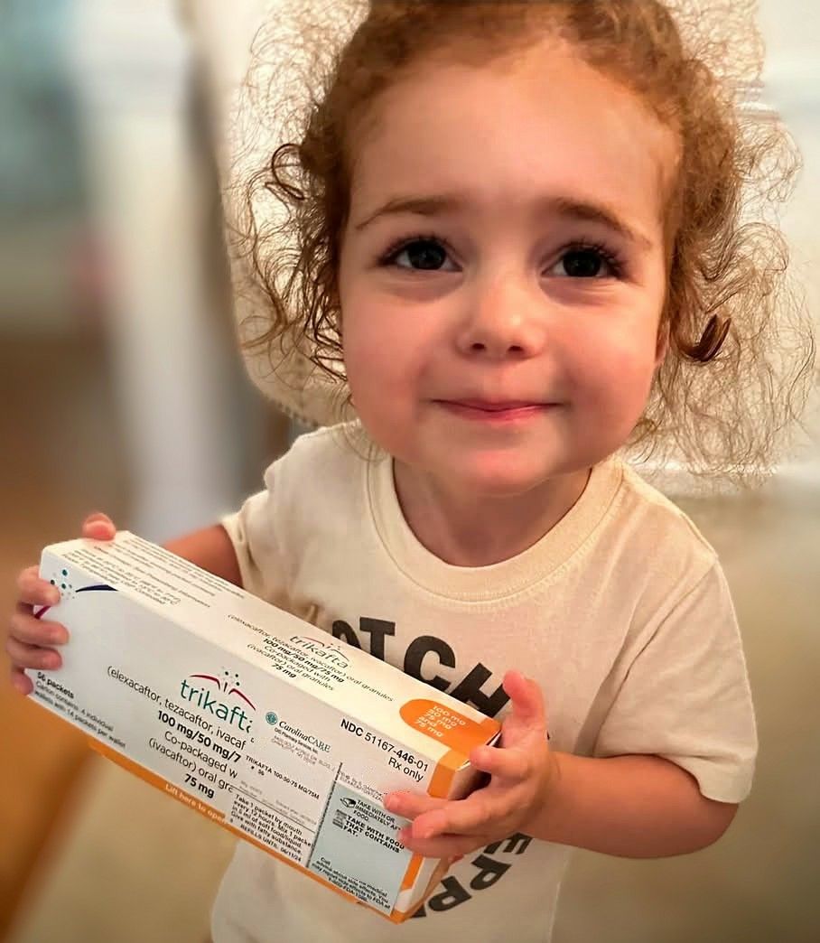 Toddler with curly red hair holding her first box of Trikafta, smiling at her mother who is off camera, but was crying with relief.