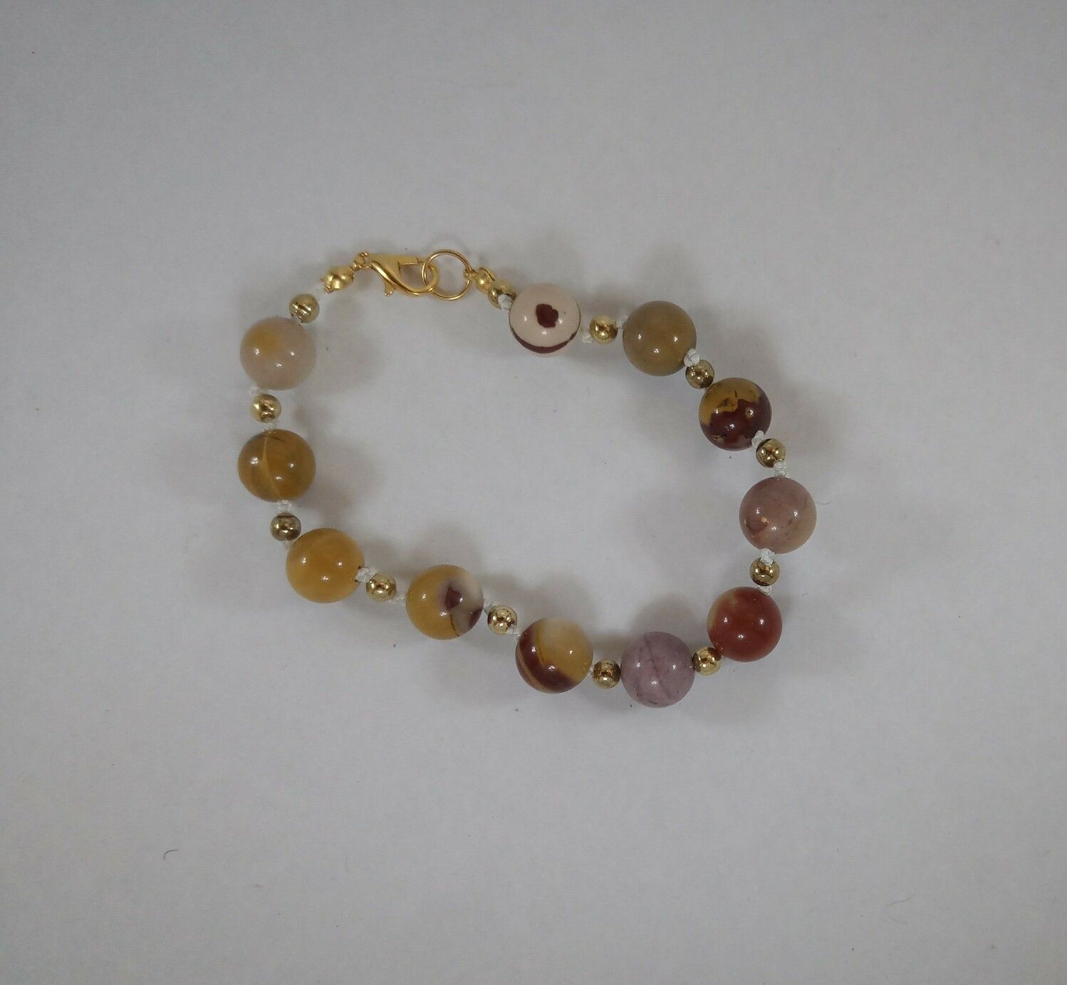 A mookaite bracelet, strung on white nylon cord, with gold spacer beads. Mookaite is a fossil stone, and it comes in a lot of colours that are sometimes banded or swirled - this bracelet is mostly orange, red and brown shades, but there's one bead that's cream with swirls of dark brown, and another that's almost a Lavender purple.